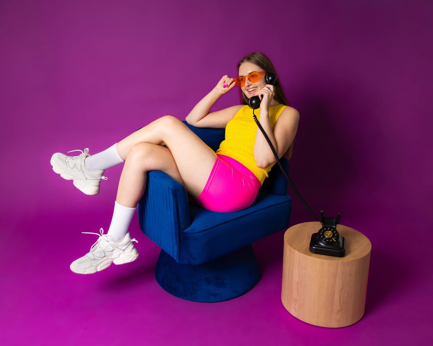 Young woman in yellow top and pink shorts talks on retro phone, lounging in blue chair against purple background.