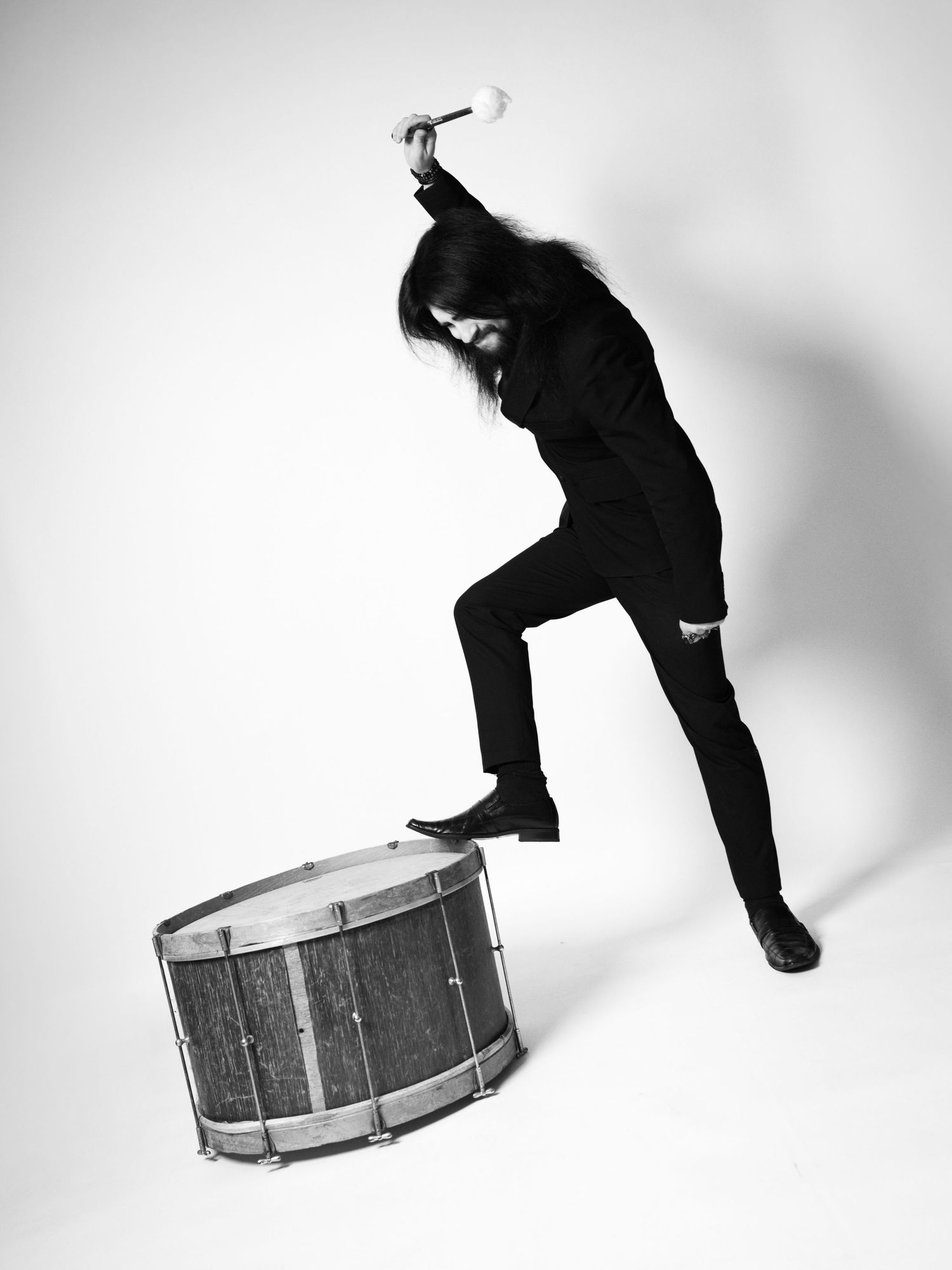 Black and white photo of a long-haired musician in black clothes raising a drumstick over a snare drum.