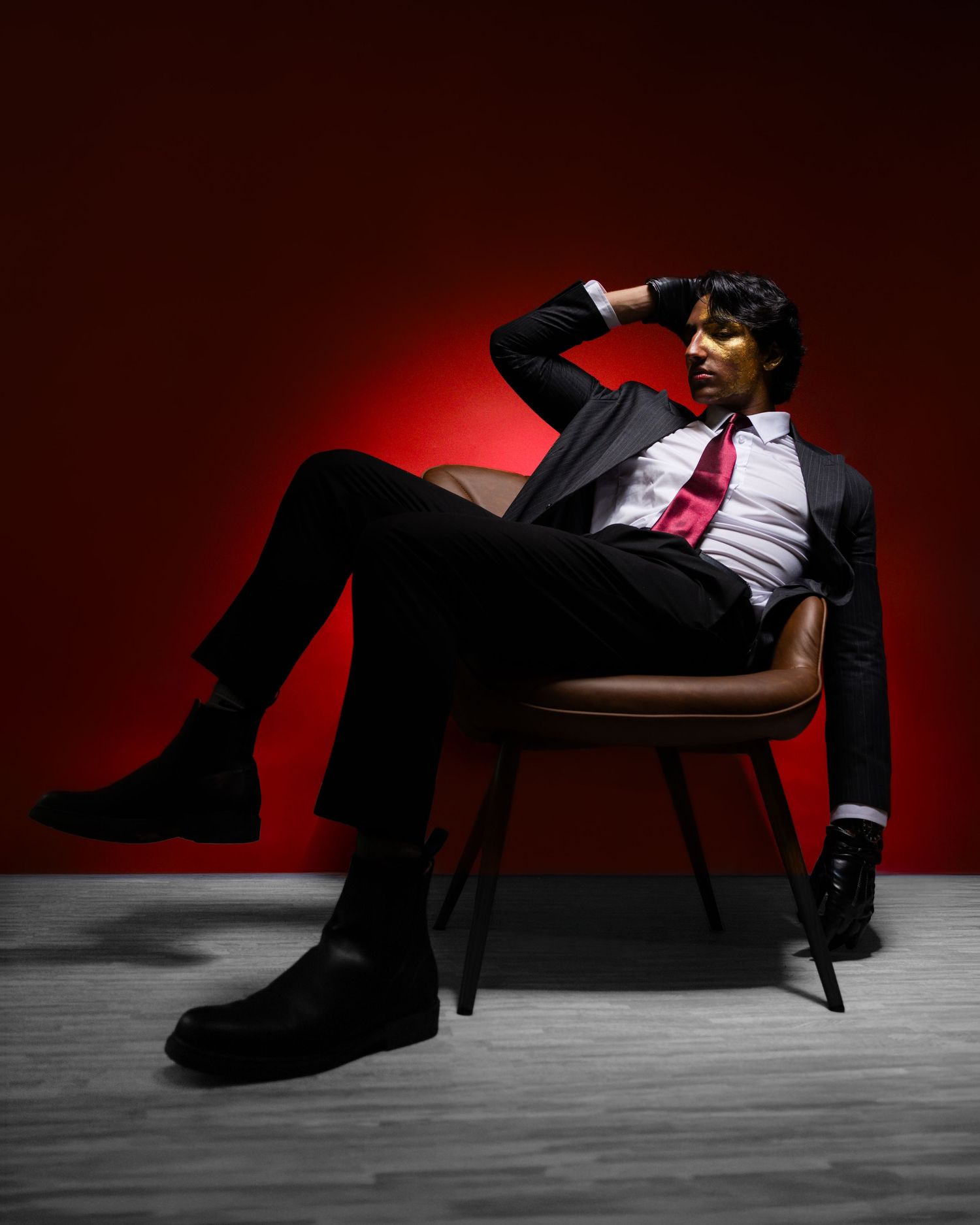 A stylish man in a black suit and red tie sits relaxed in a chair against a dramatic red background.
