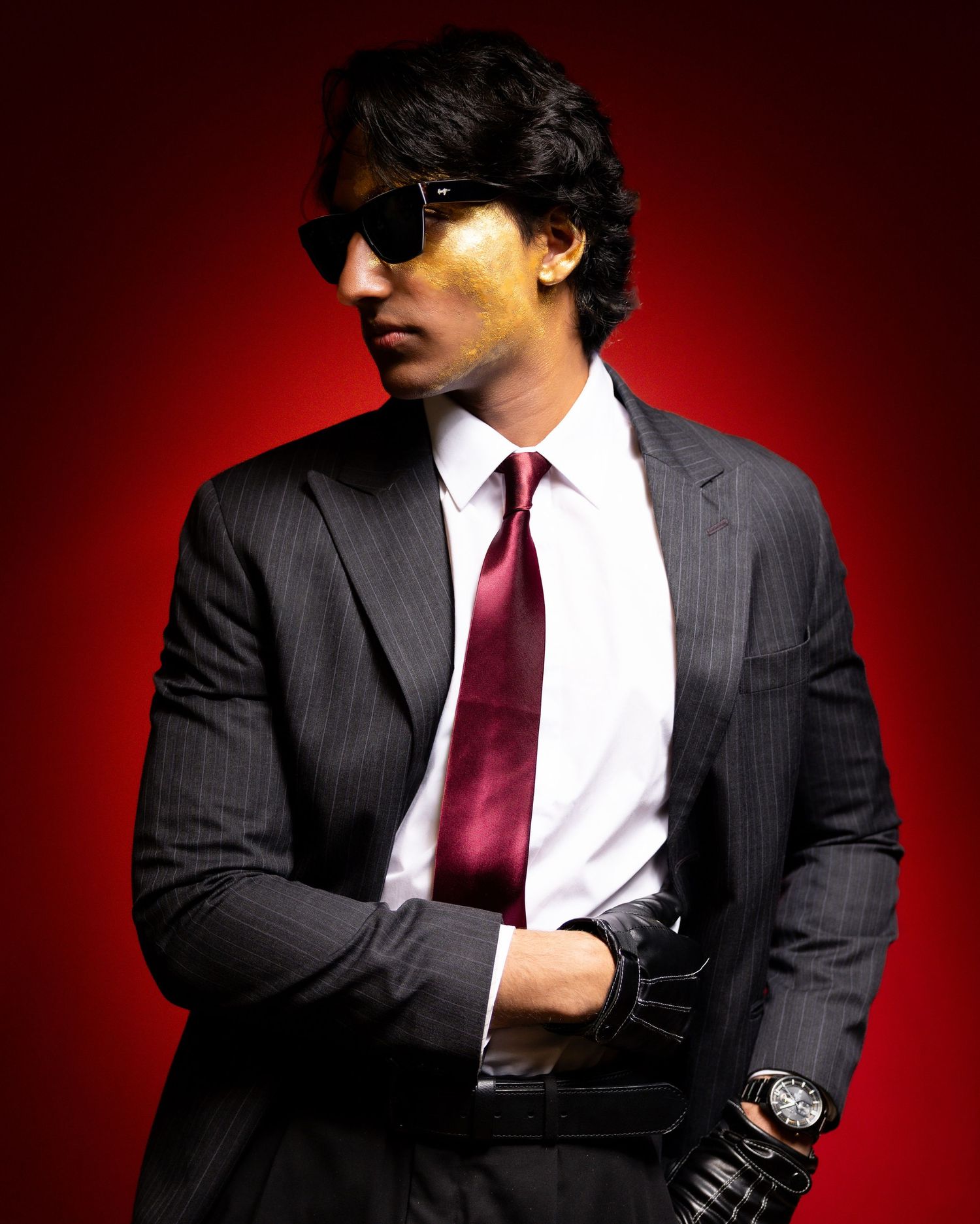 Stylish man in pinstripe suit with red tie, sunglasses, and leather gloves posing against red background.