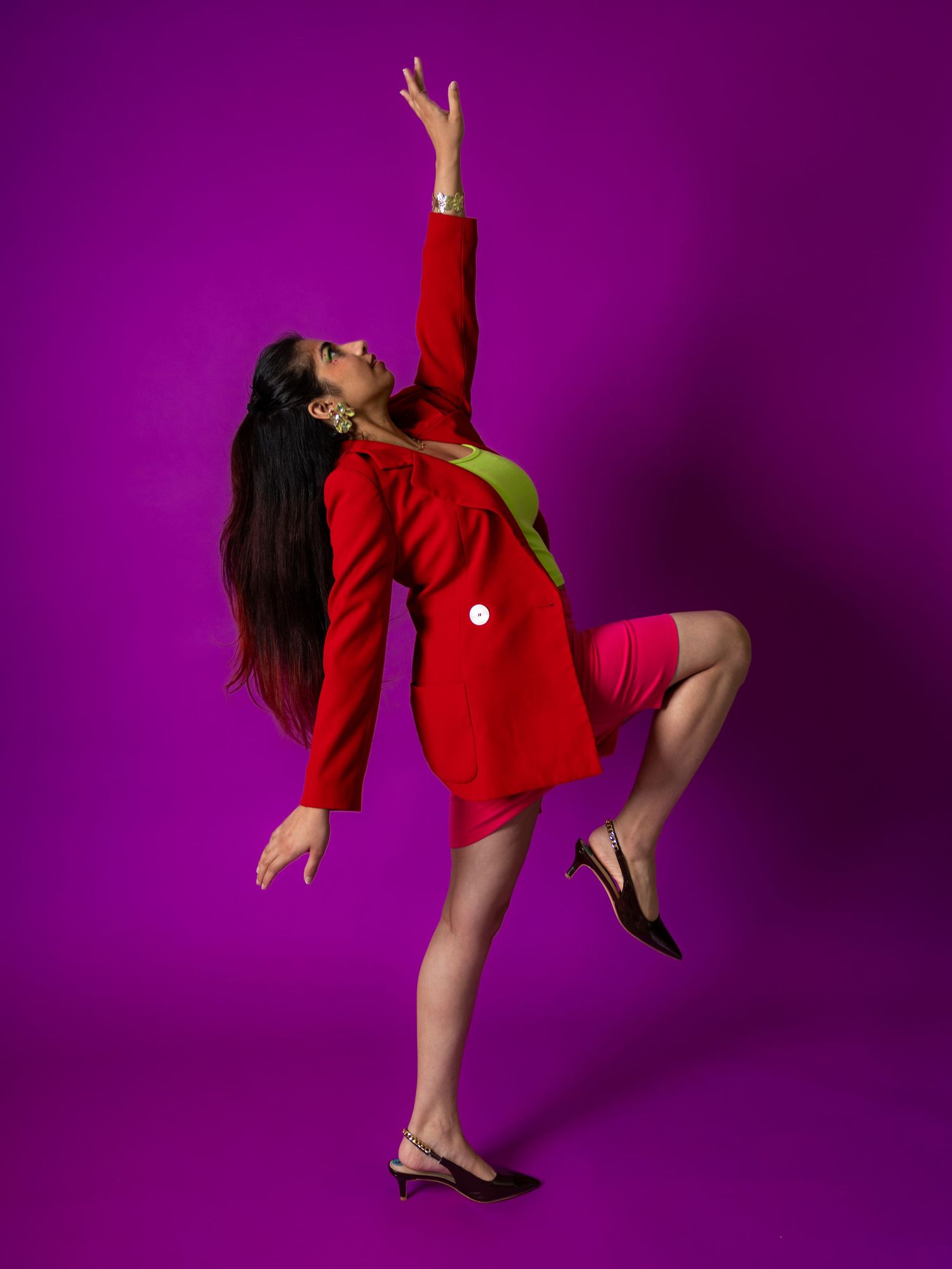 Woman in red blazer and pink skirt poses dynamically against vibrant purple background with one leg raised.