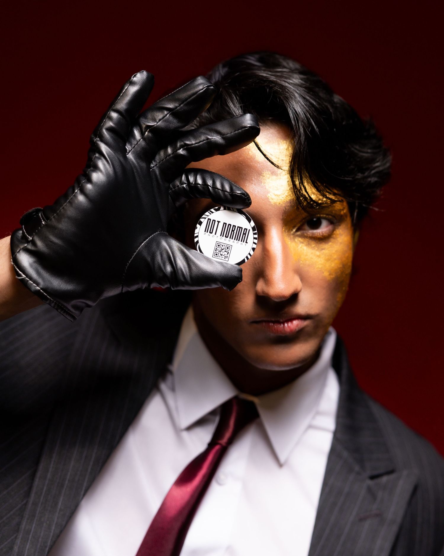 Person in suit and black glove holding a button badge over one eye against a dark red background.
