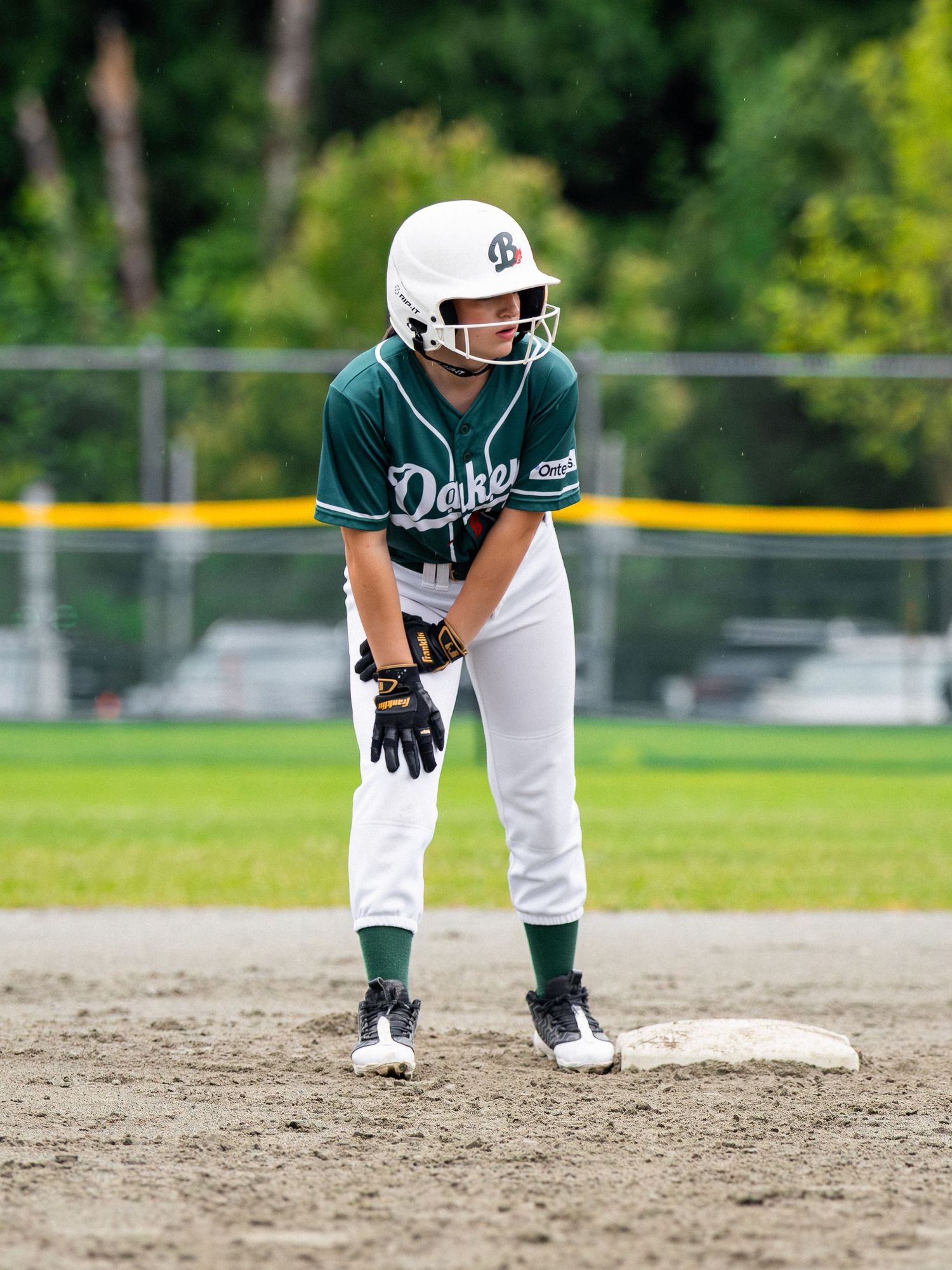 Youth softball player in green Oakes jersey and white helmet stands ready at base on a dirt infield.