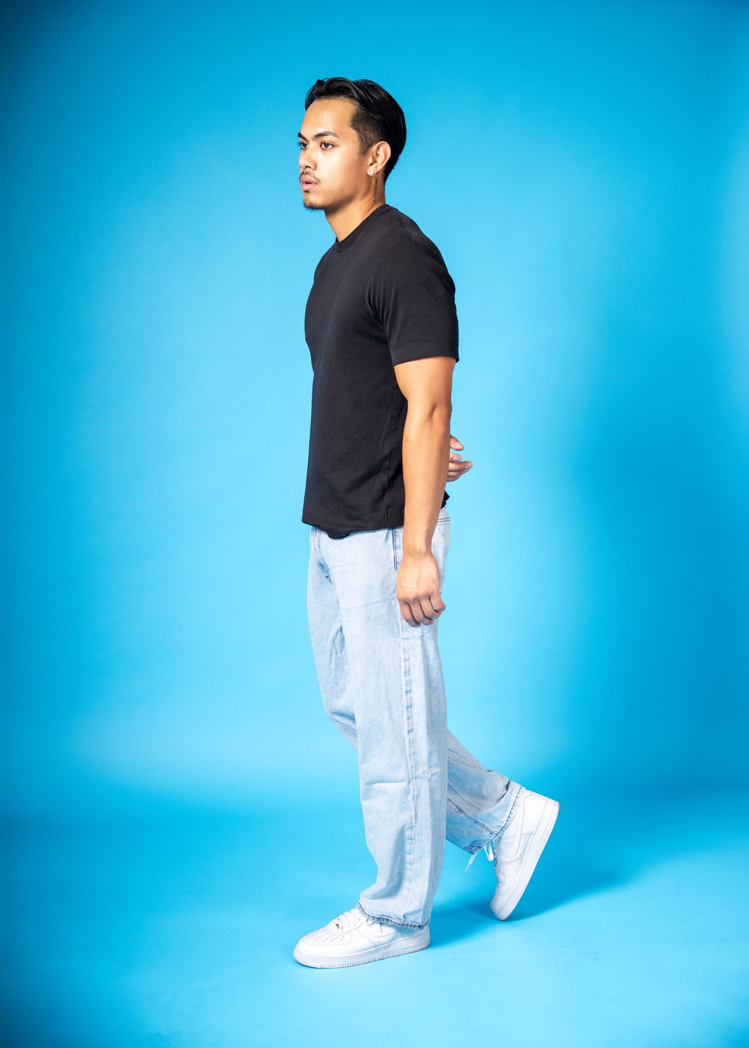 Man in black t-shirt, light wash jeans, and white sneakers posing sideways against a blue background.