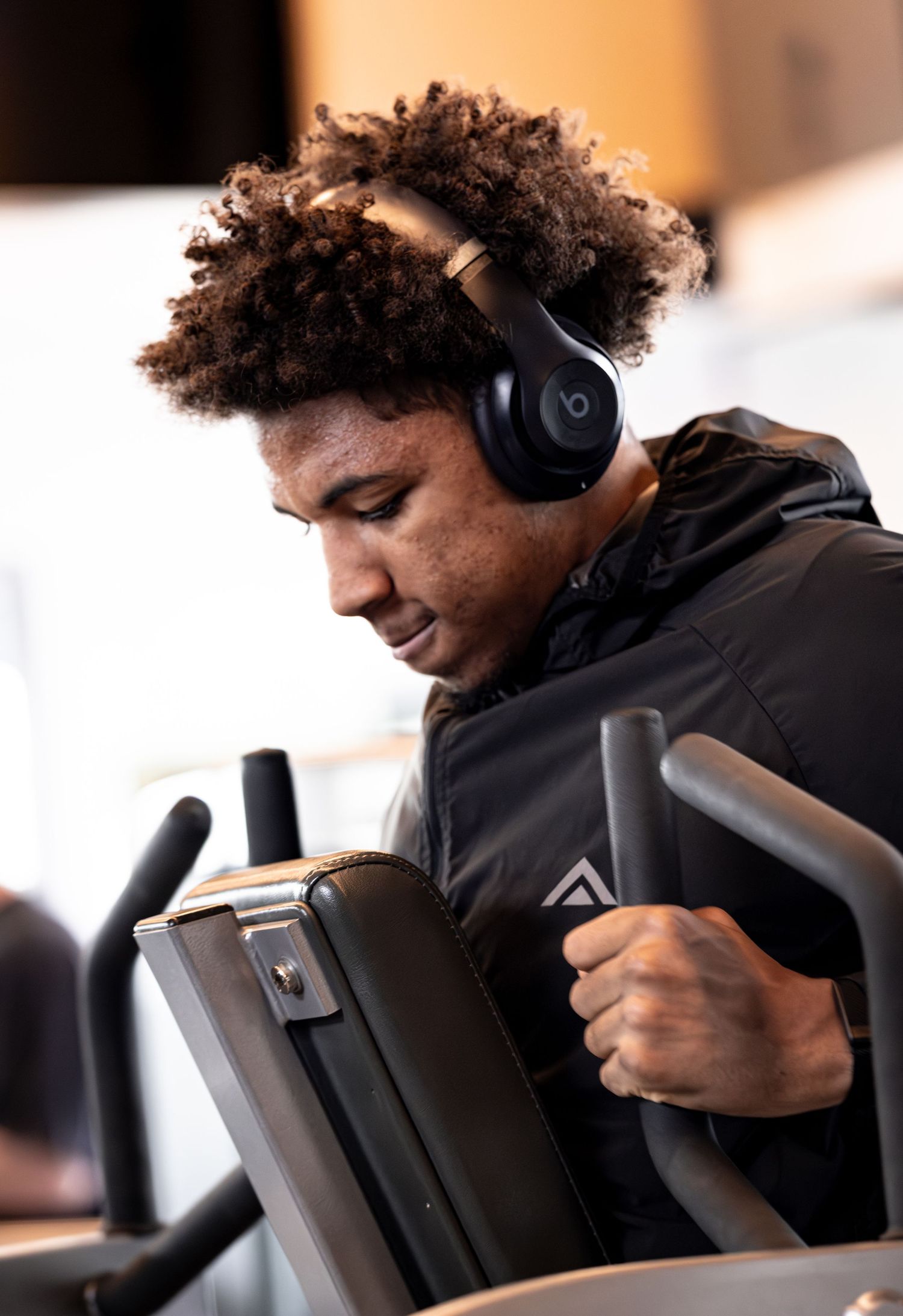 Young man with curly hair wearing Beats headphones works out on an elliptical machine at the gym.