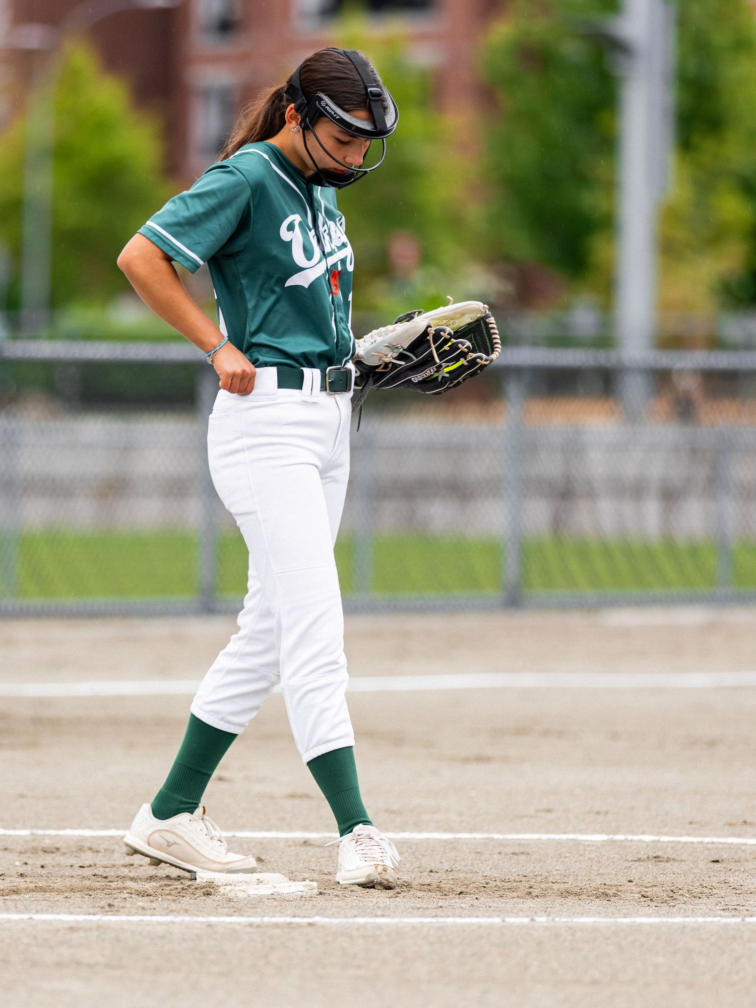 Female softball player in green uniform walking on field holding glove during outdoor game.