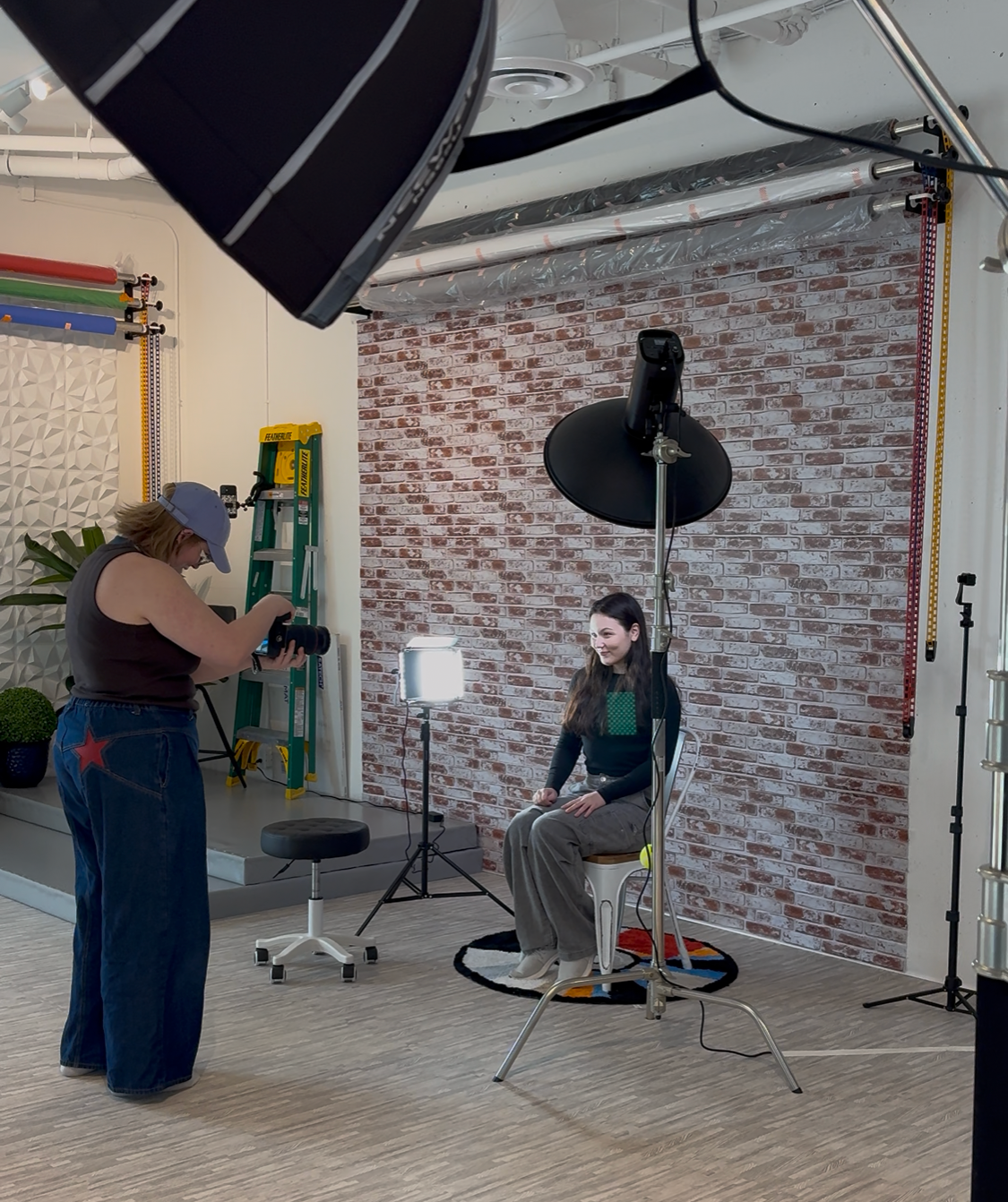 Photographer taking professional studio portrait of woman seated on chair with brick backdrop and lighting equipment.