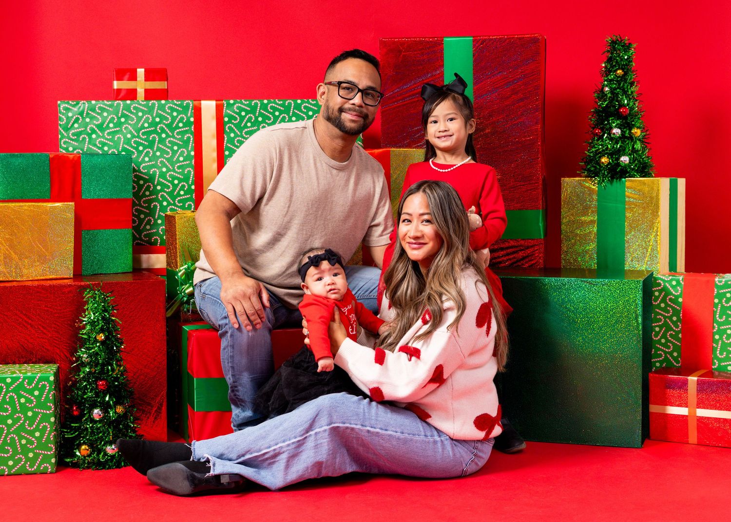 Family of four poses joyfully among colorful Christmas gifts and mini trees on a festive red background.