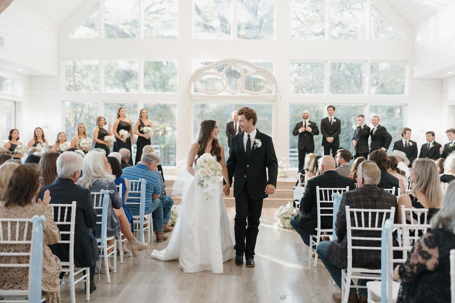 A wedding ceremony in progress inside an elegant glass-walled venue with white chairs and natural light streaming in.