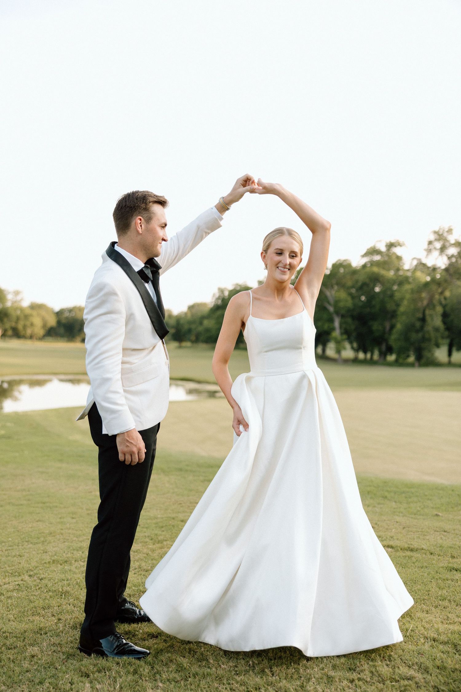 A couple dances together on a lush green lawn with trees and a pond in the background during a wedding celebration.