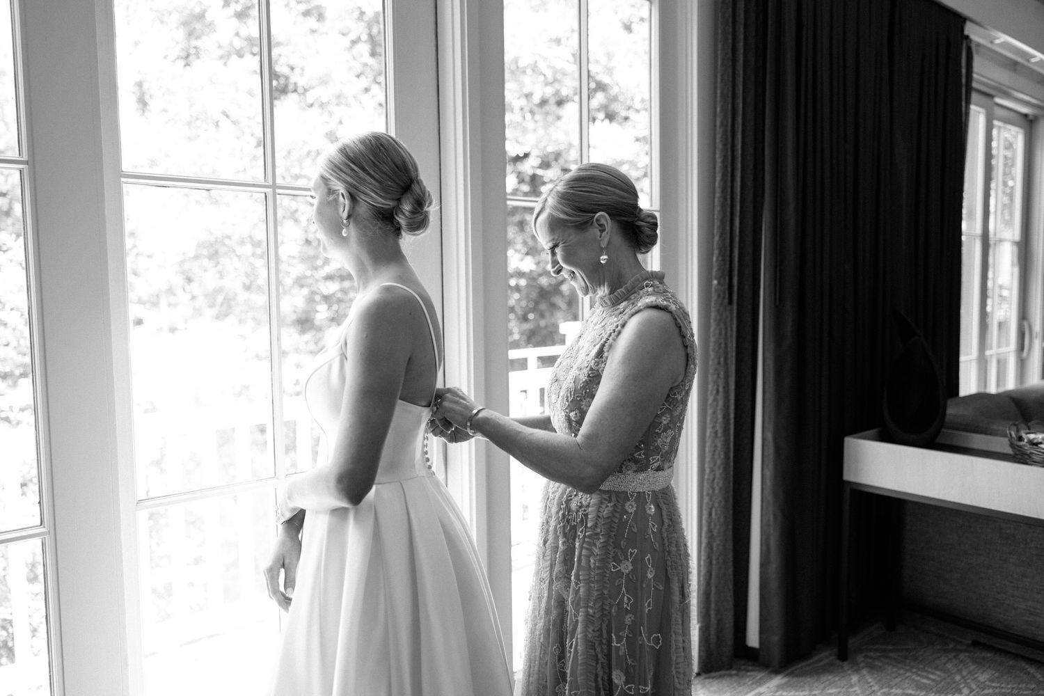 A tender moment captured in black and white as a bride gets help with her dress near a bright window.