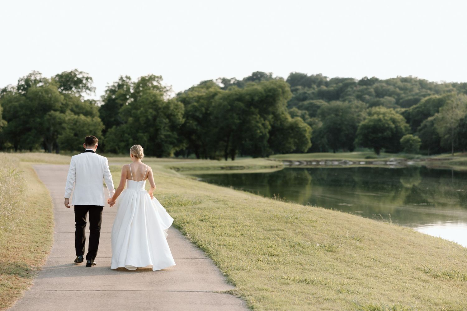 A couple in wedding attire walks along a peaceful lakeside path surrounded by green trees on a sunny day.