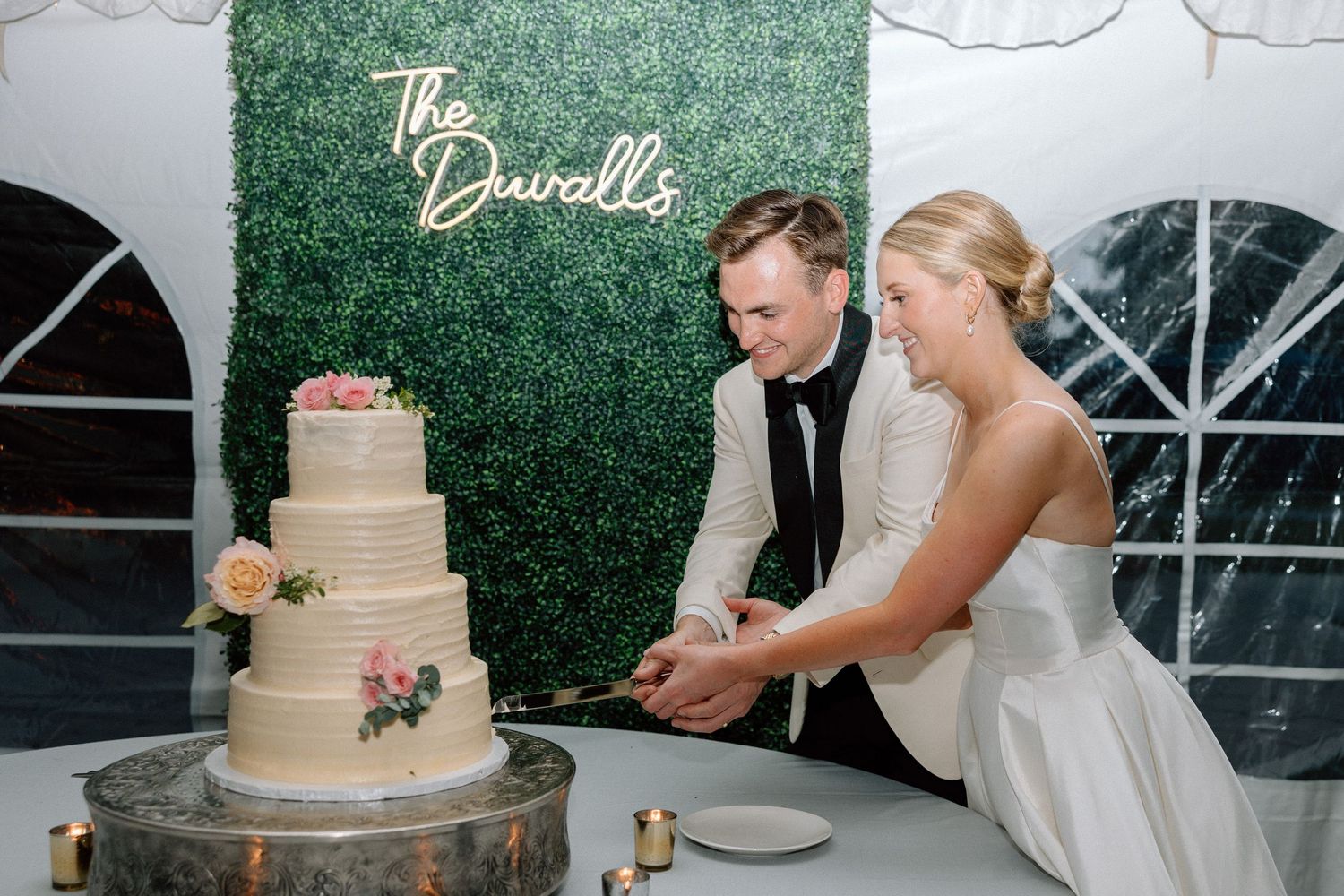 A couple cuts their elegant three-tiered wedding cake adorned with blush roses against a hedge wall backdrop.