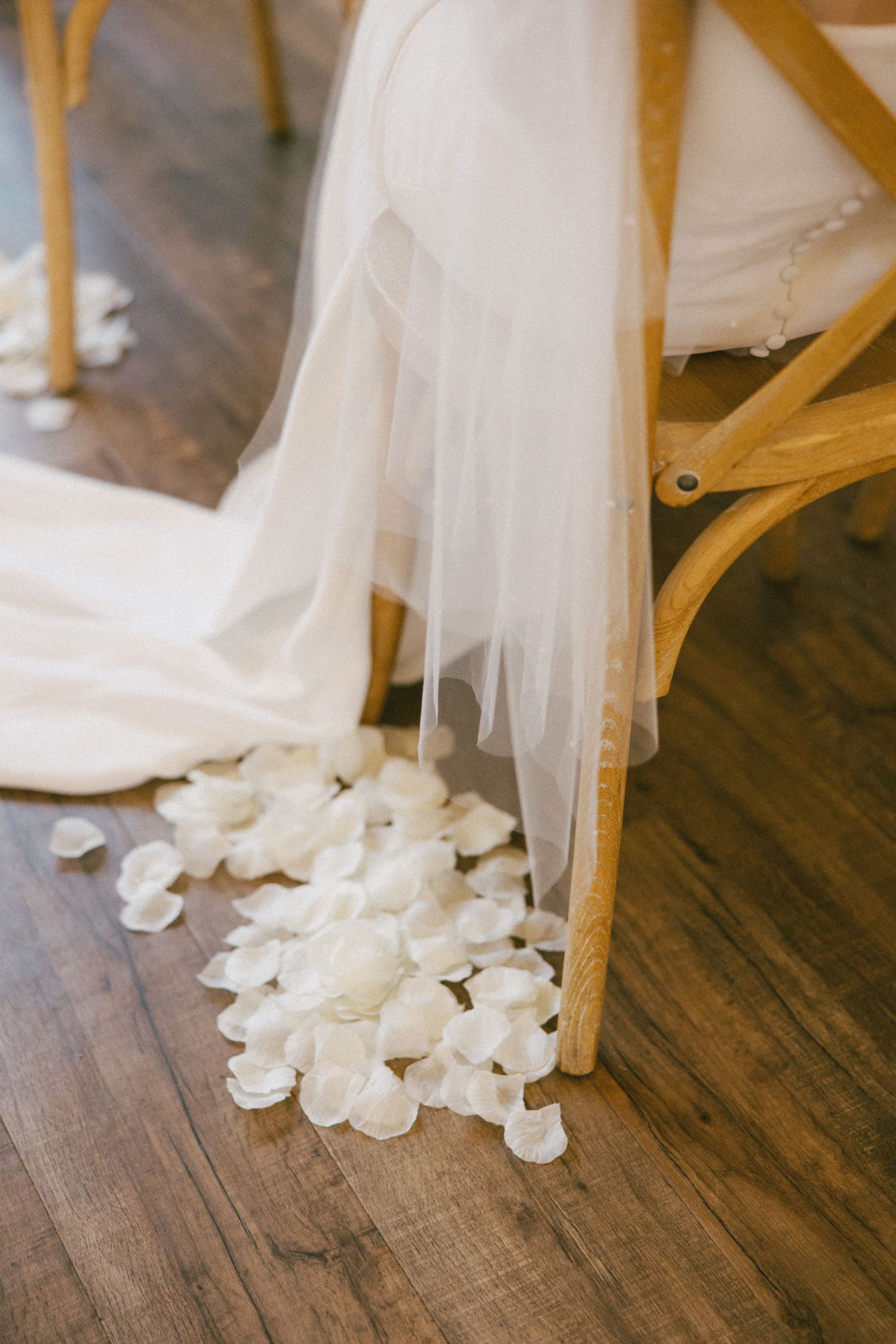 White rose petals scattered on hardwood floor beneath flowing wedding dress and wooden chair.