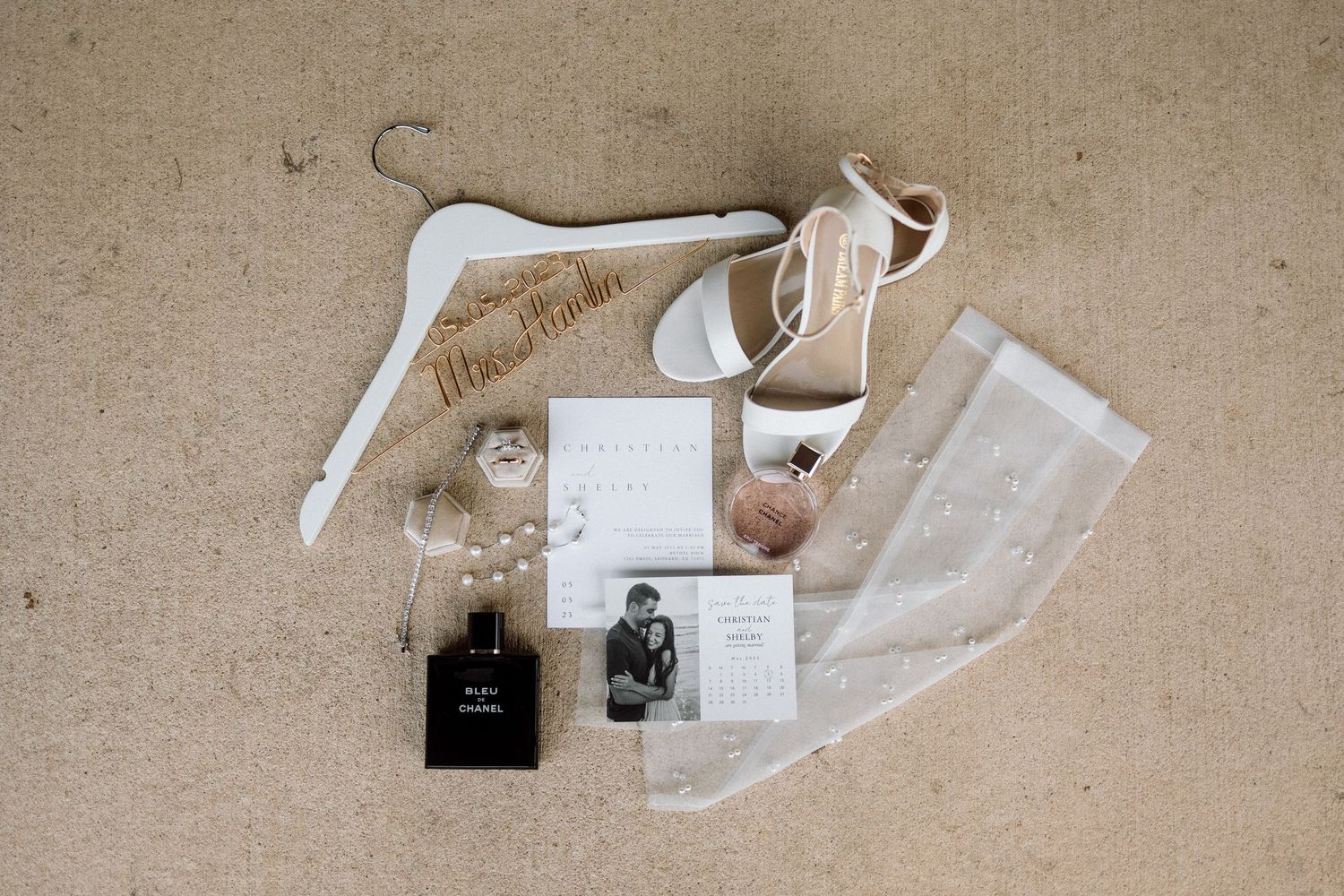 Wedding invitation suite with white ribbon, cards, and envelopes arranged on a beige textured background.