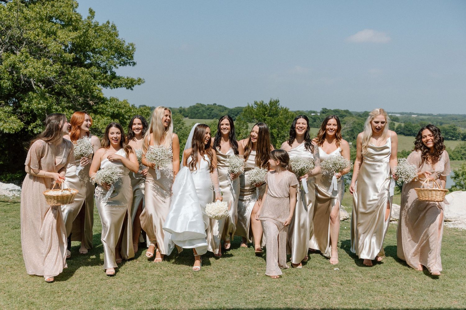 Wedding party in light pink dresses walks together across a sunny green field on a summer afternoon.