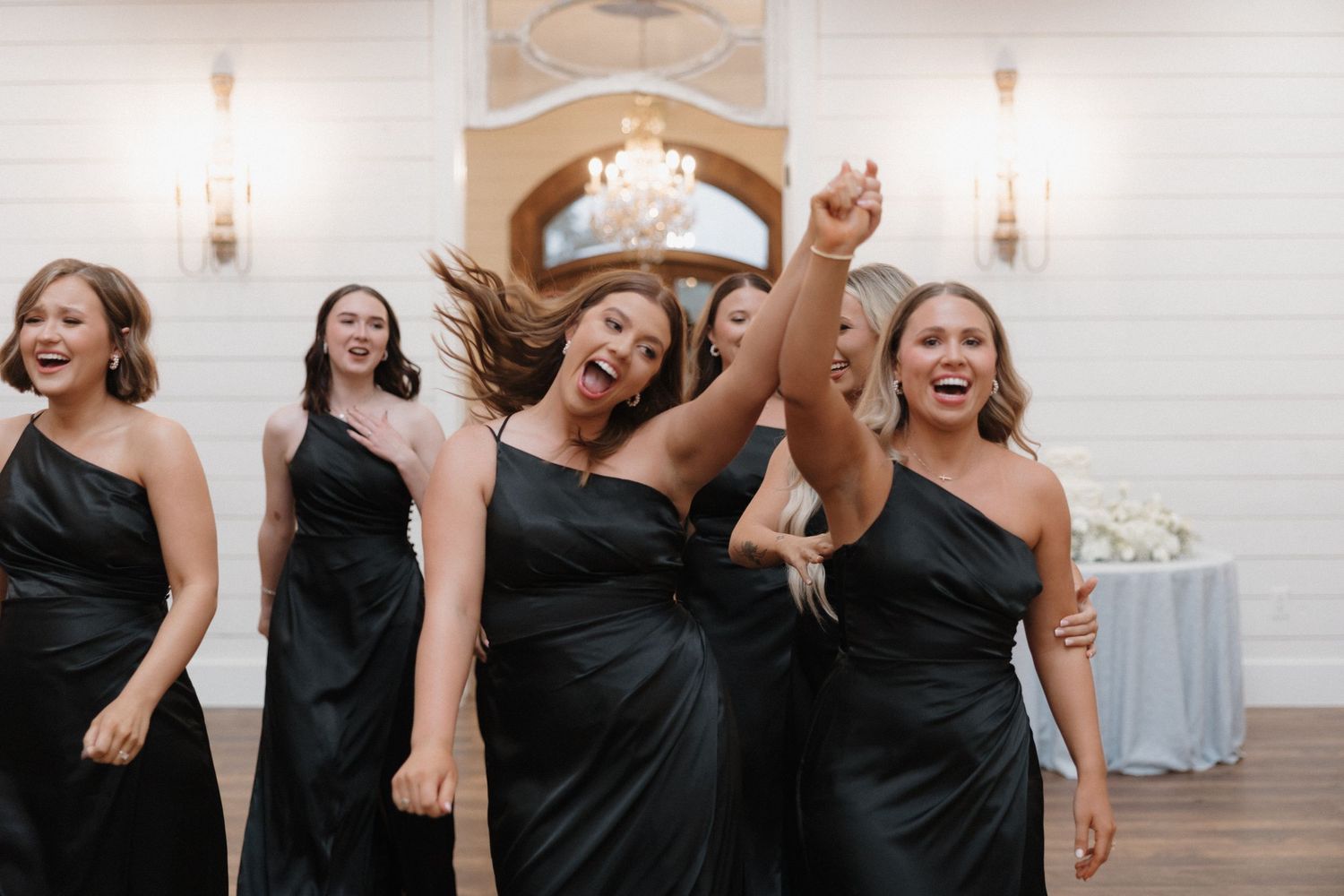 Bridesmaids in matching black one-shoulder dresses dance and celebrate joyfully at an elegant reception venue.