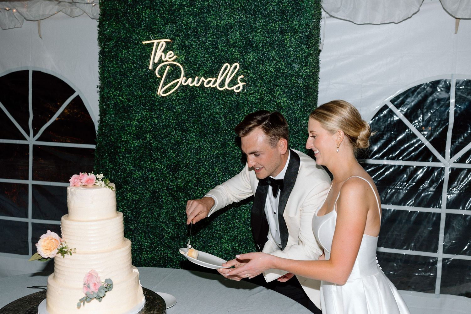 A couple cuts their elegant white wedding cake together in front of a green hedge wall backdrop at their reception.