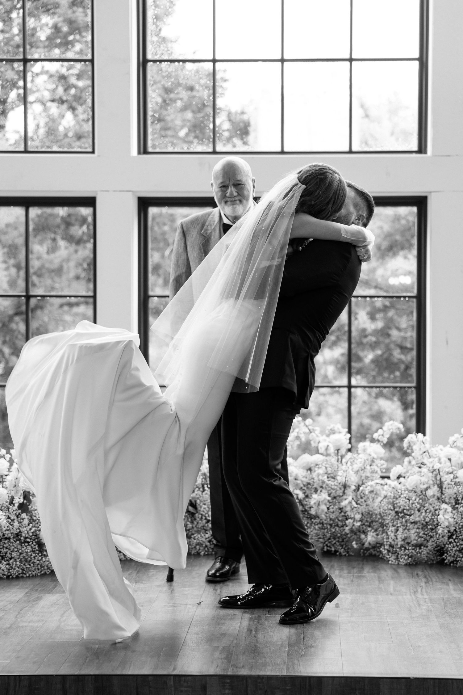 A couple in formal attire performs a dramatic dip dance move in front of large windows in a bright room.