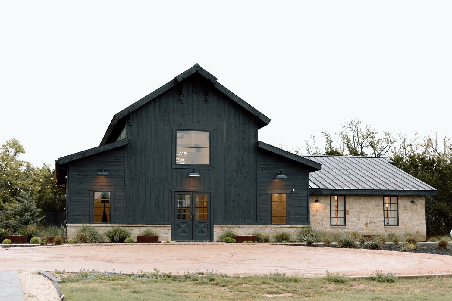 A modern dark-colored barn building with multiple sections and a metal roof stands against a cloudy sky.