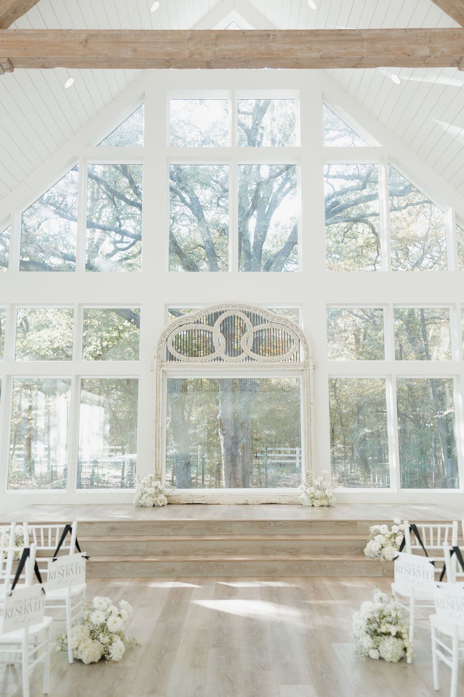 Elegant white conservatory wedding venue with floor-to-ceiling windows, rustic wood beams, and floral arrangements.