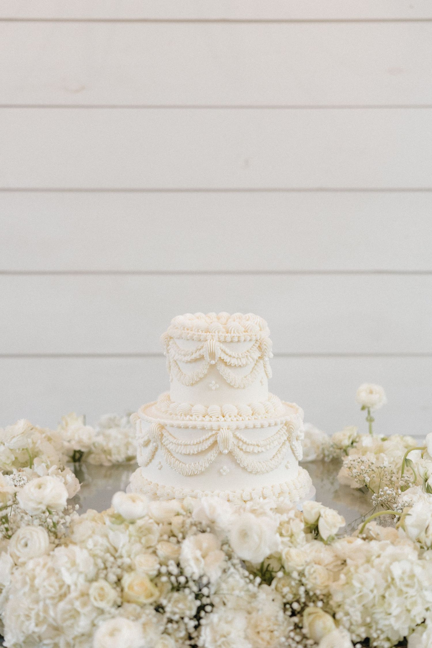 Elegant white wedding cake with pearl details sits atop a bed of lush white hydrangeas and roses against a gray backdrop.