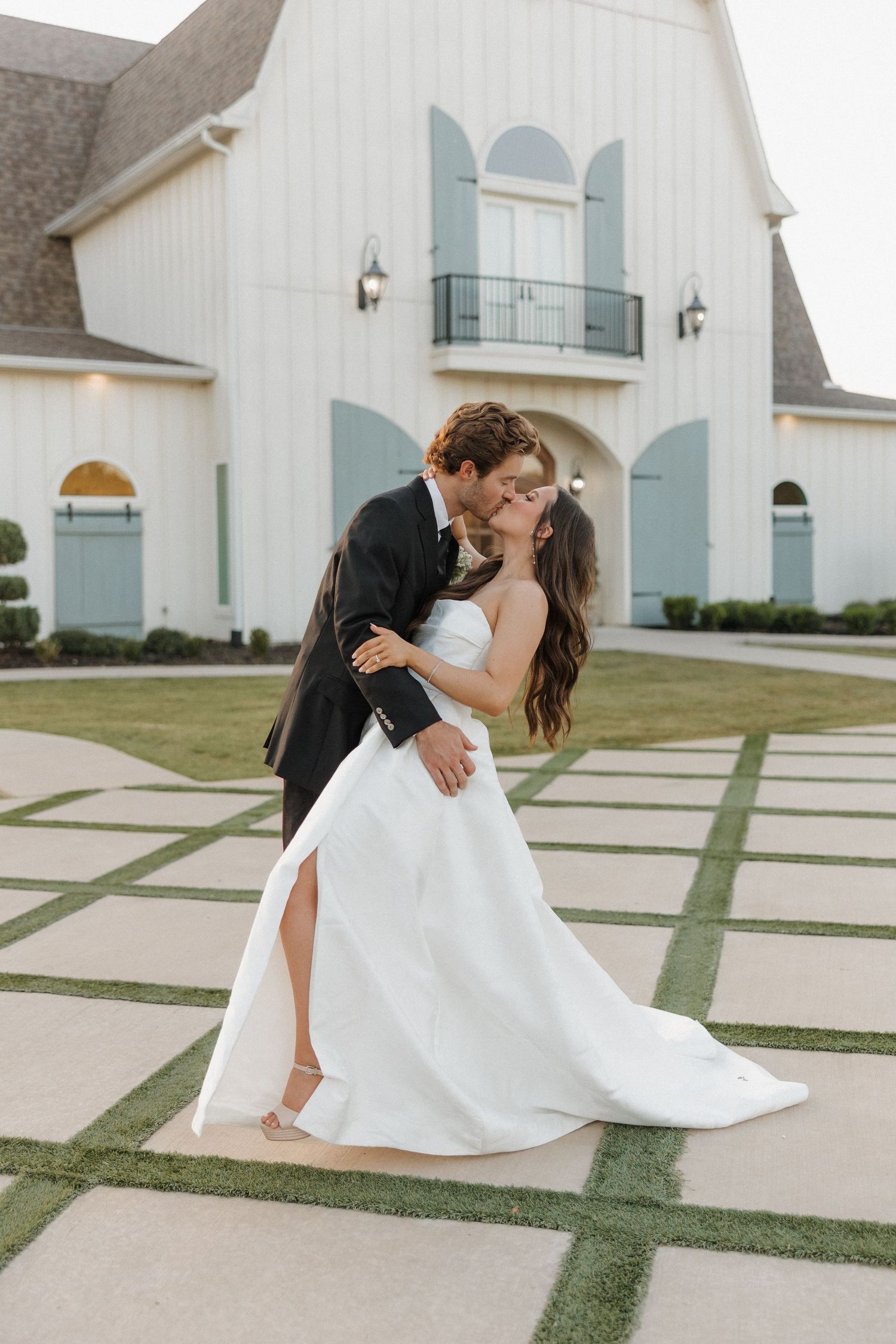 A romantic couple shares a tender kiss on a paved pathway in front of a white church building.