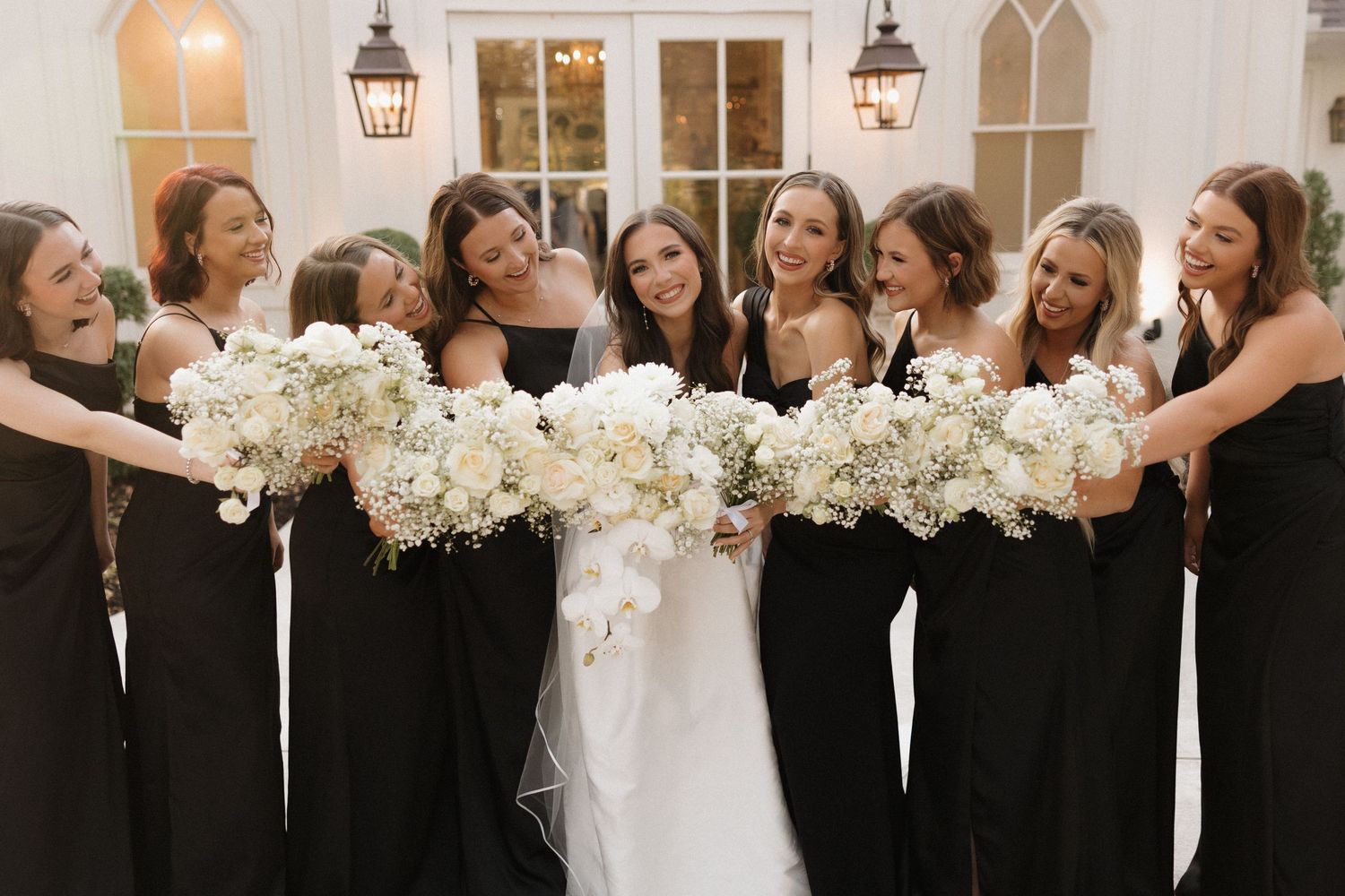 Elegant wedding party in black attire poses with white floral bouquets in front of a bright church window.