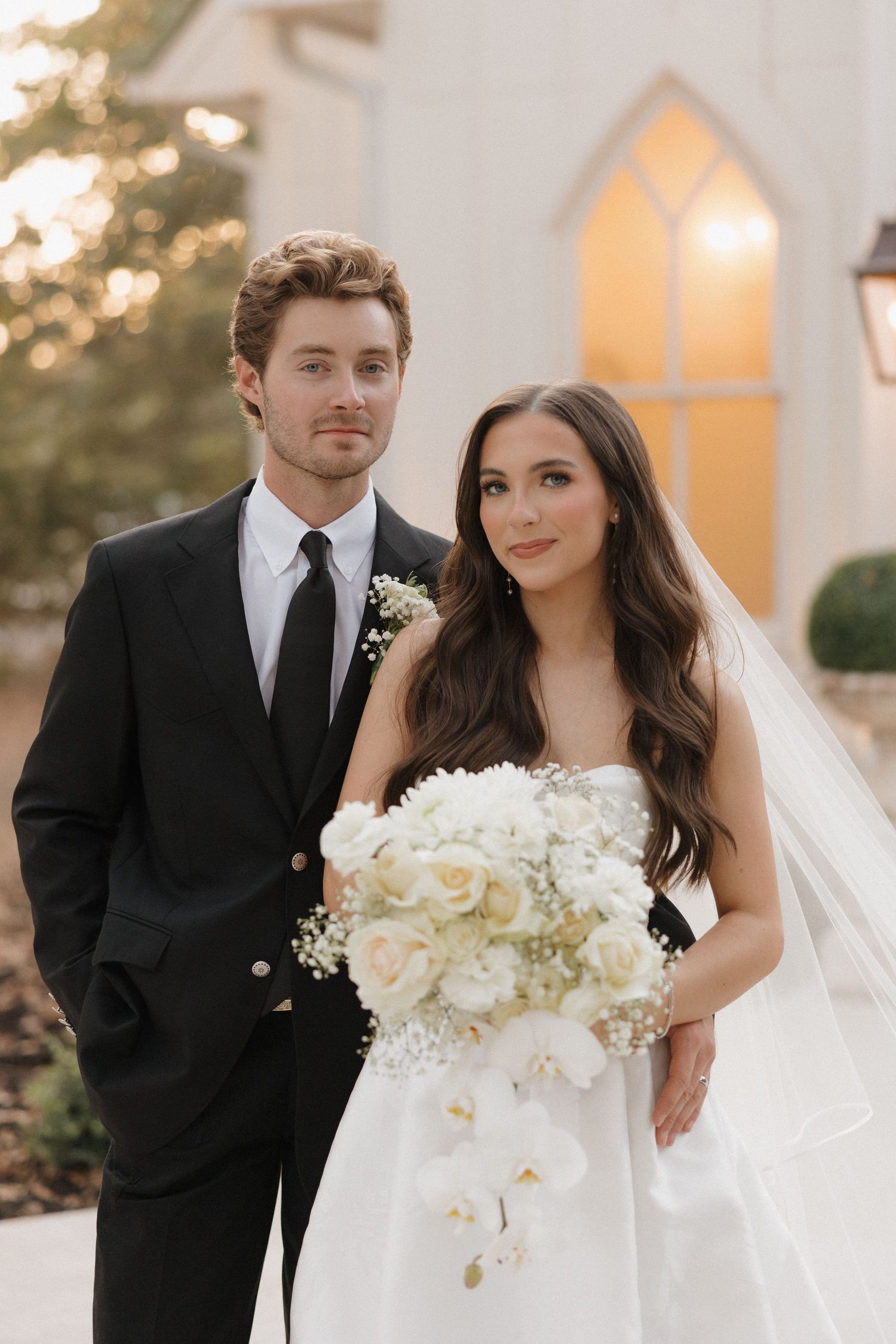 A bride and groom share intimate wedding portraits outdoors in front of a white chapel at sunset.