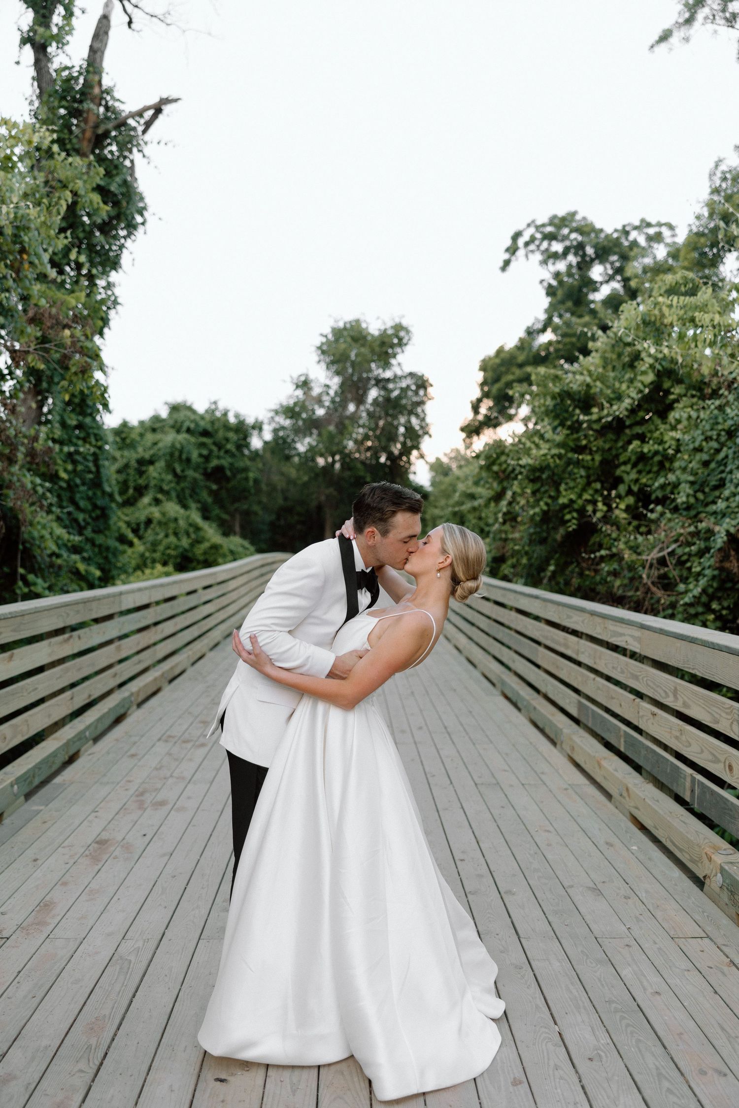 A couple in wedding attire share a romantic kiss on a wooden deck surrounded by lush green trees.