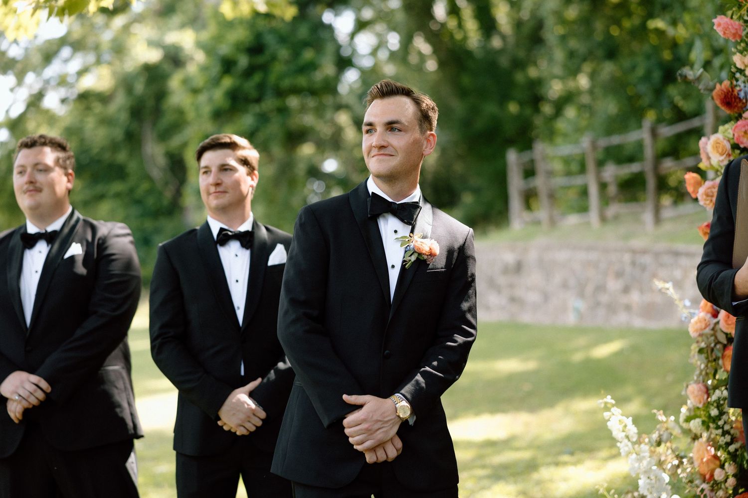 Groomsmen in black tuxedos pose together on a sunny day outdoors near stone wall and greenery.