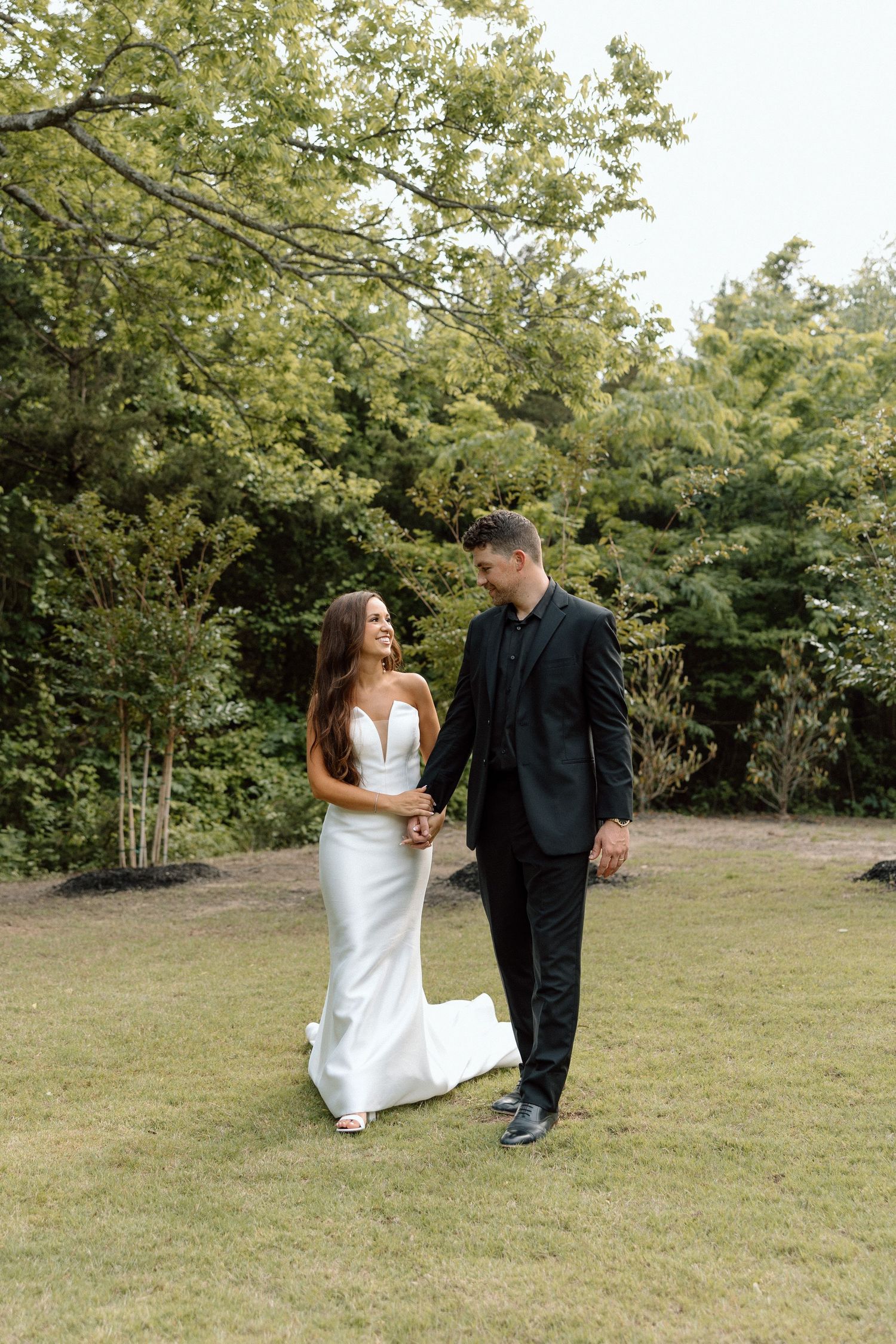 A couple shares a candid moment together in a grassy field surrounded by trees during their wedding photoshoot.