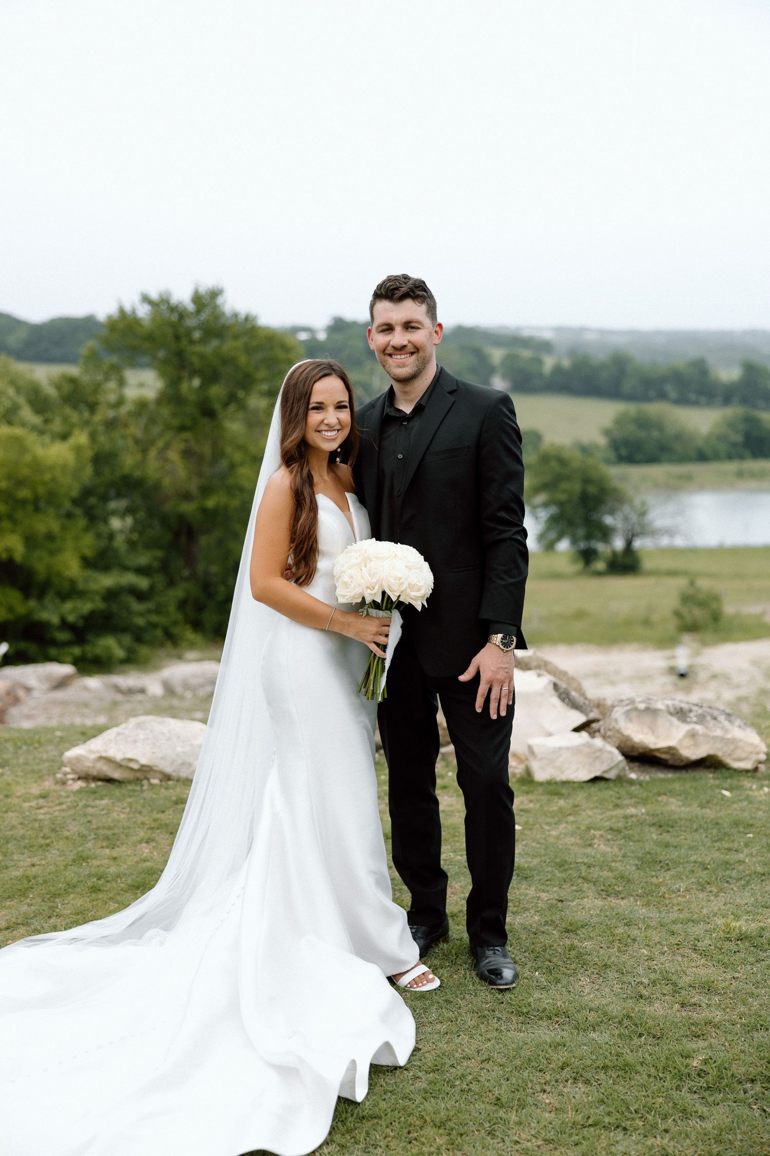 A couple poses outdoors on their wedding day with a scenic lake and rolling hills in the background.