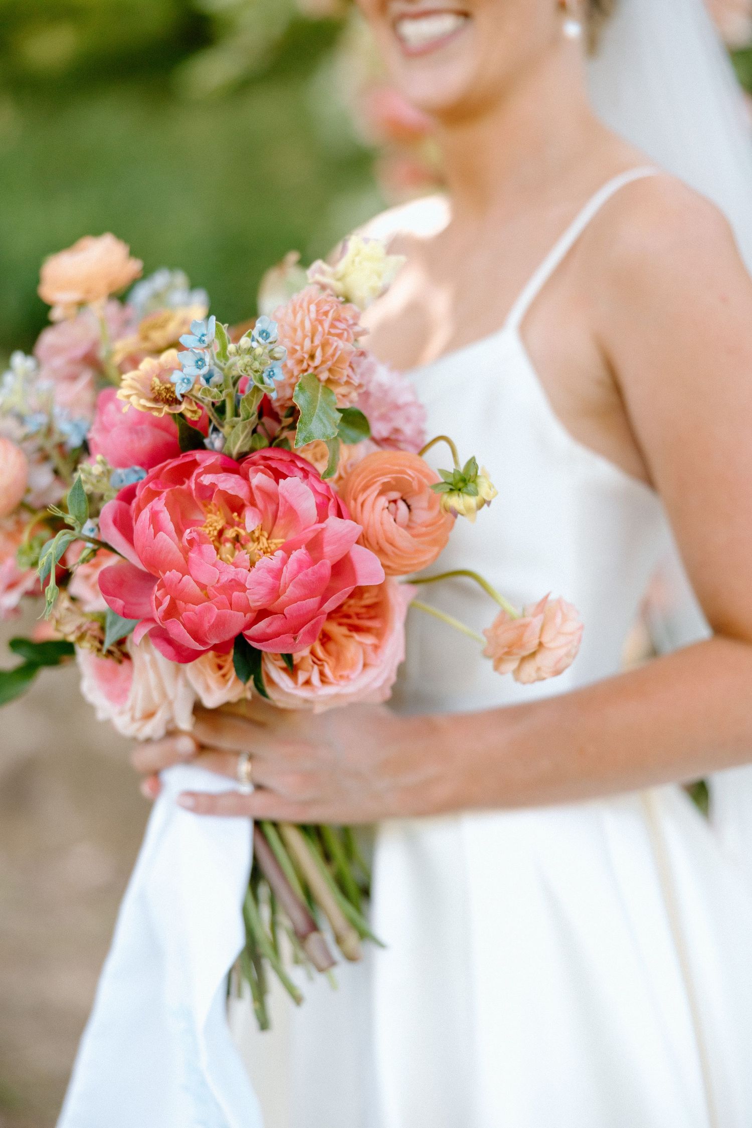 A vibrant coral and peach bridal bouquet with peonies and ranunculus against a white wedding dress.