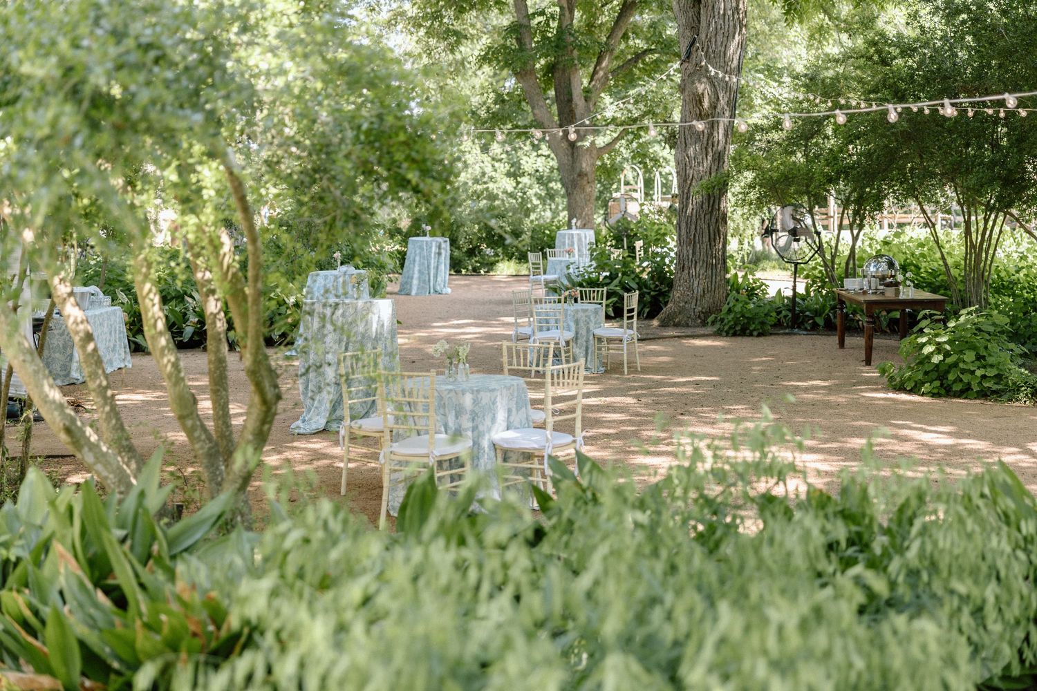 White chairs arranged in a garden setting with dappled sunlight filtering through trees for an outdoor ceremony.