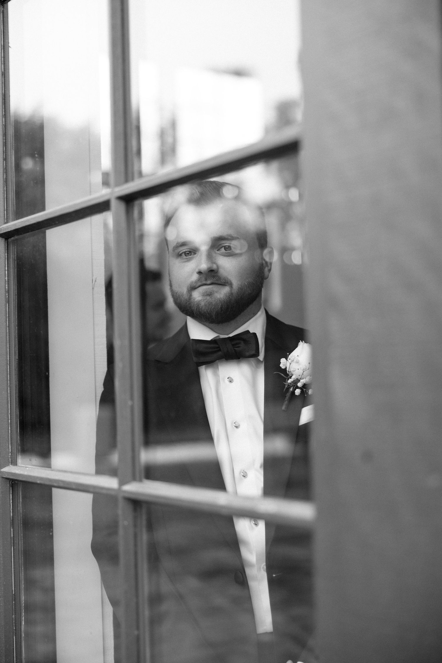 Black and white photo showing a reflection of a groom in formal attire through a window pane.