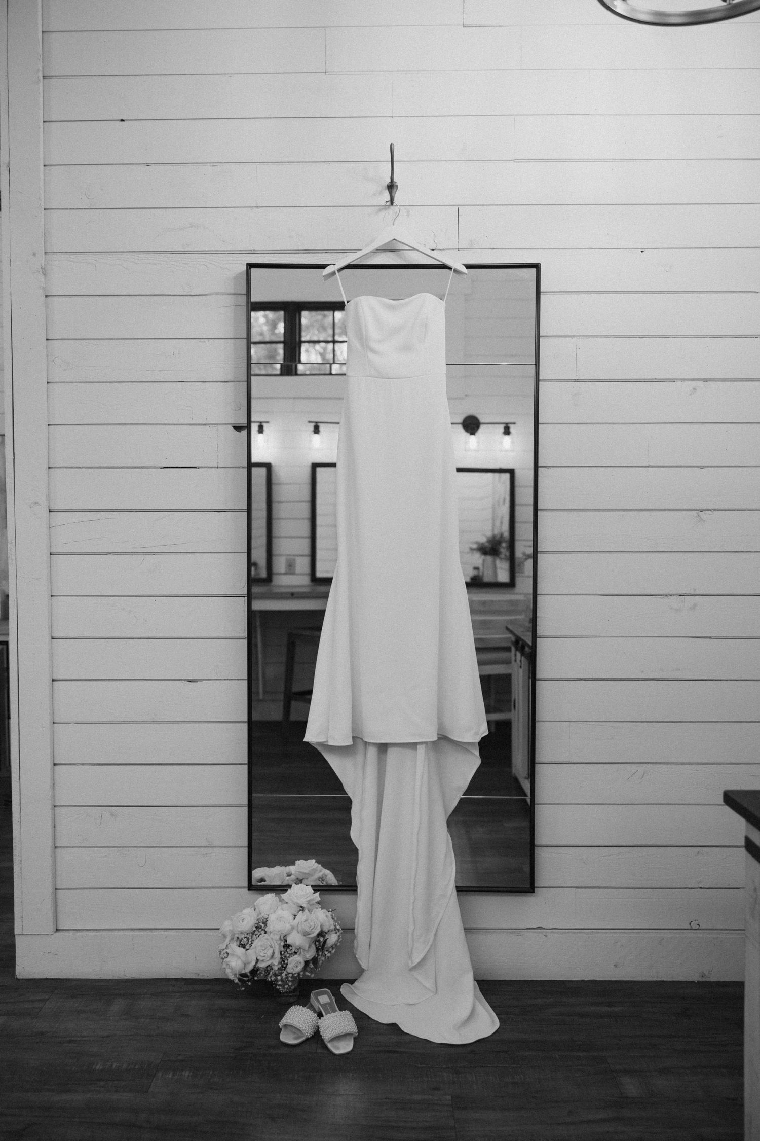 A white wedding dress hangs on a mirror against a white wooden wall, with shoes and flowers placed below.