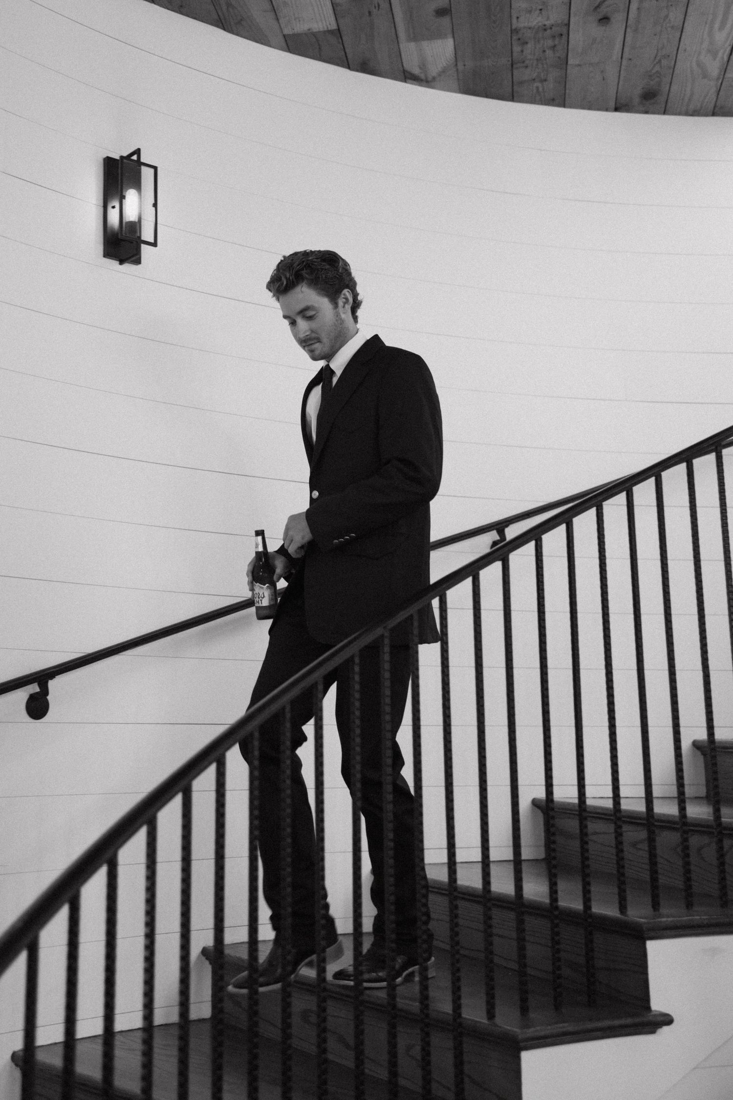Black and white photo of someone in a suit descending a curved staircase with iron railing and wall sconce lighting.