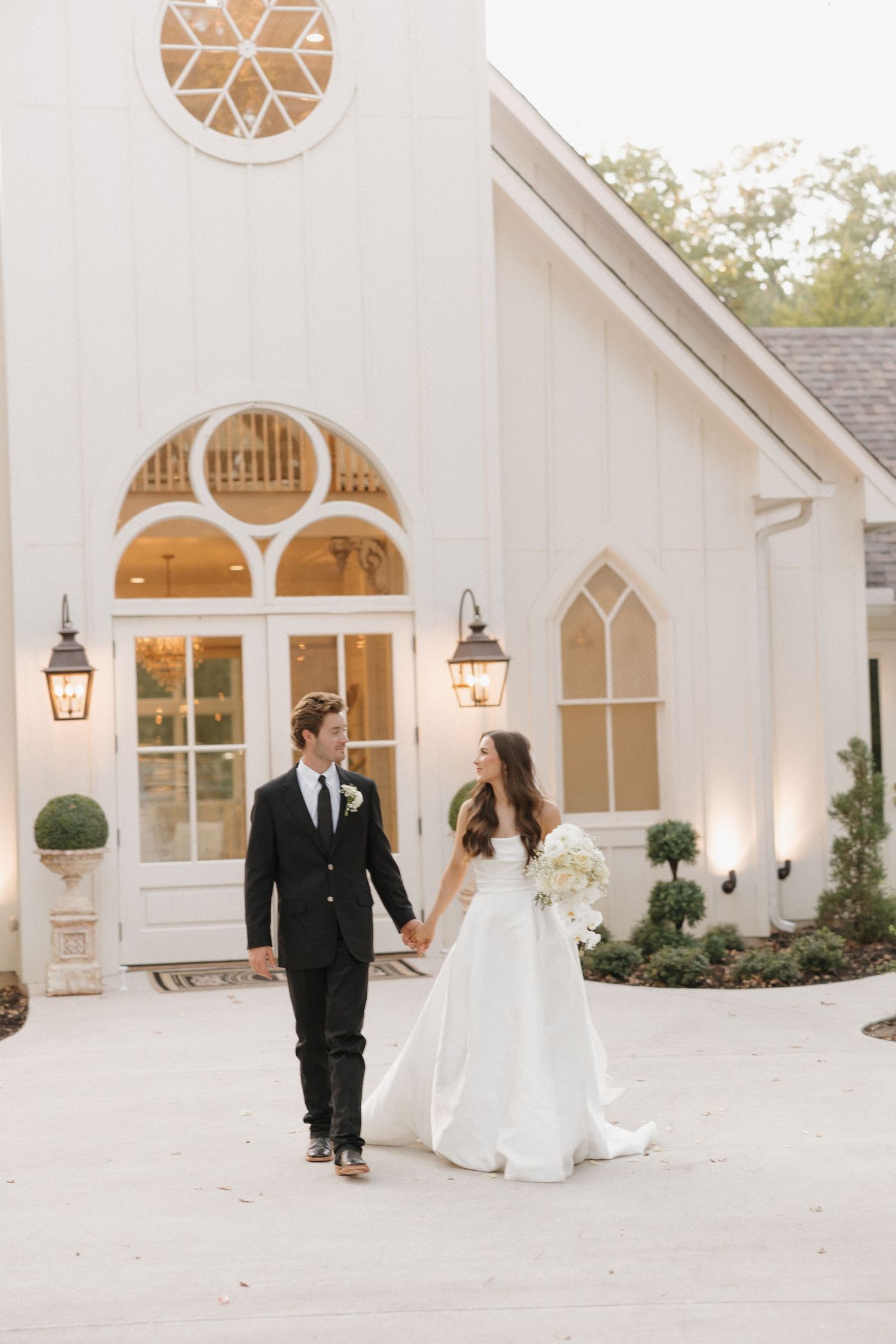 A wedding couple stands together outside a white chapel with elegant arched windows and gas lanterns.