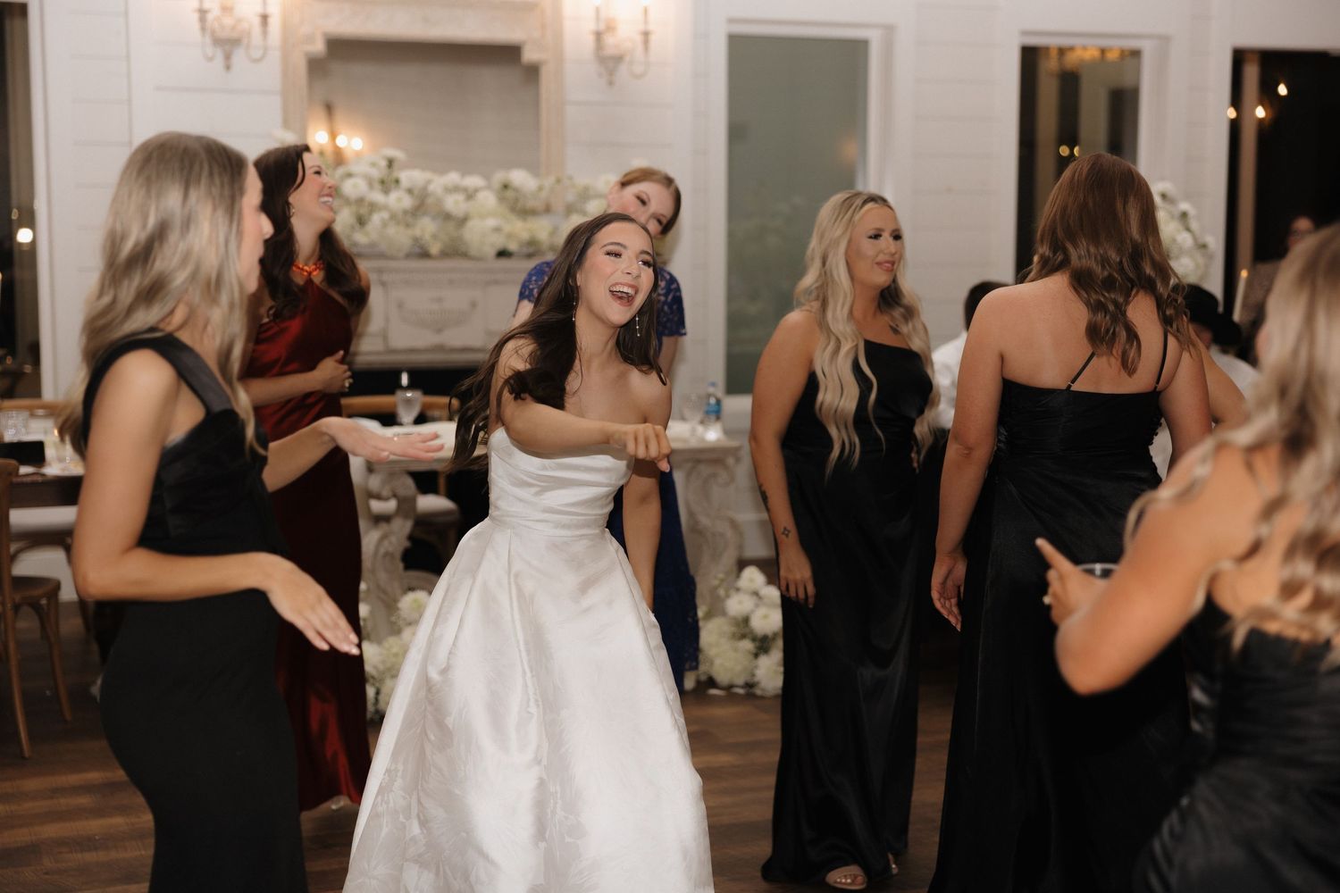 Wedding party guests in black dresses dancing and celebrating at an elegant reception venue with chandeliers.