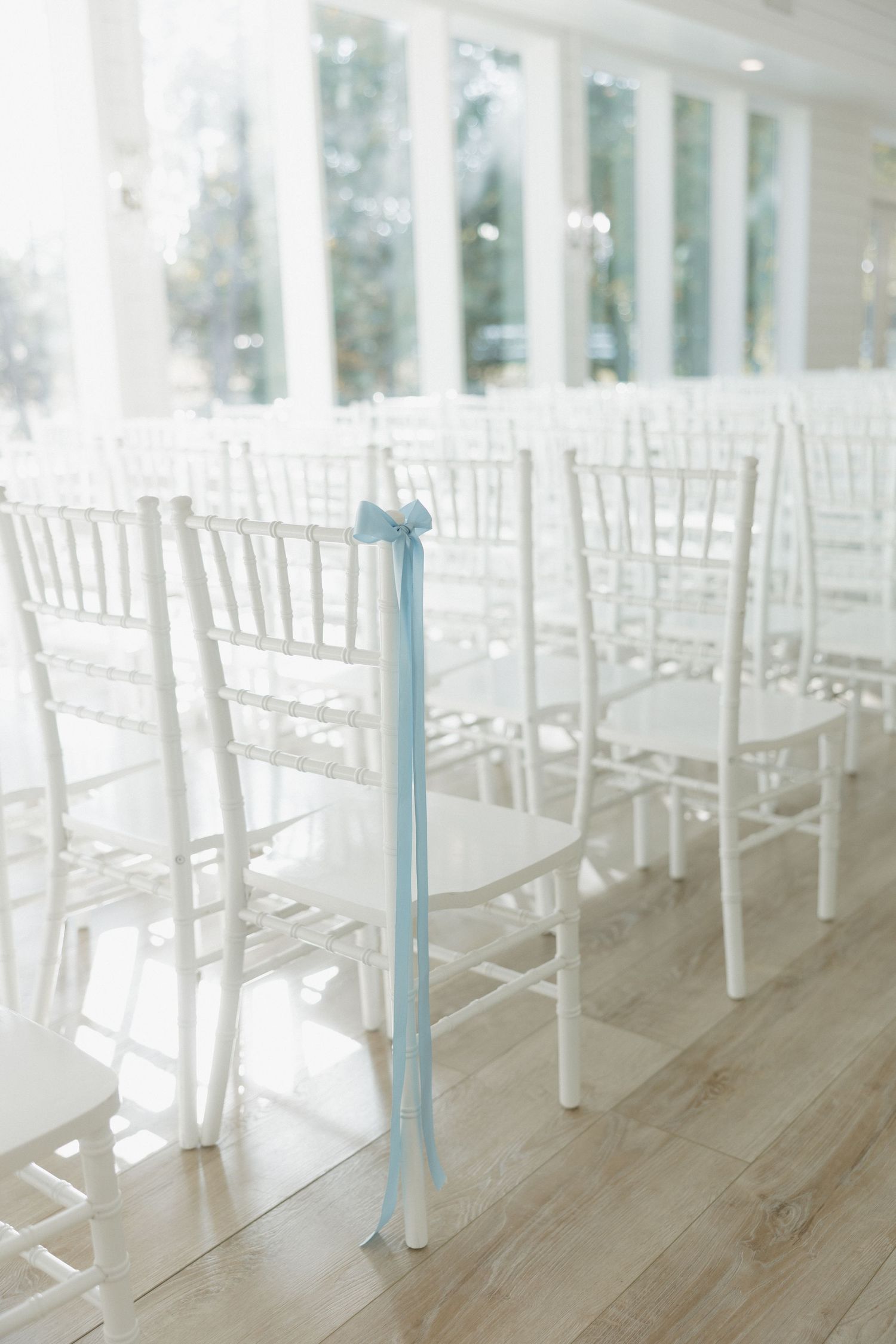 White chiavari chairs with light blue ribbon decor arranged in rows on wooden flooring in a bright indoor venue.