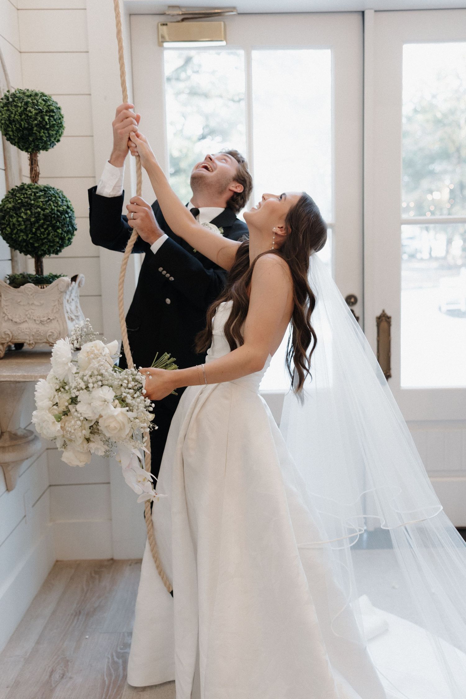 A couple in wedding attire dances joyfully in an elegant white room with decorative topiary and floral arrangements.