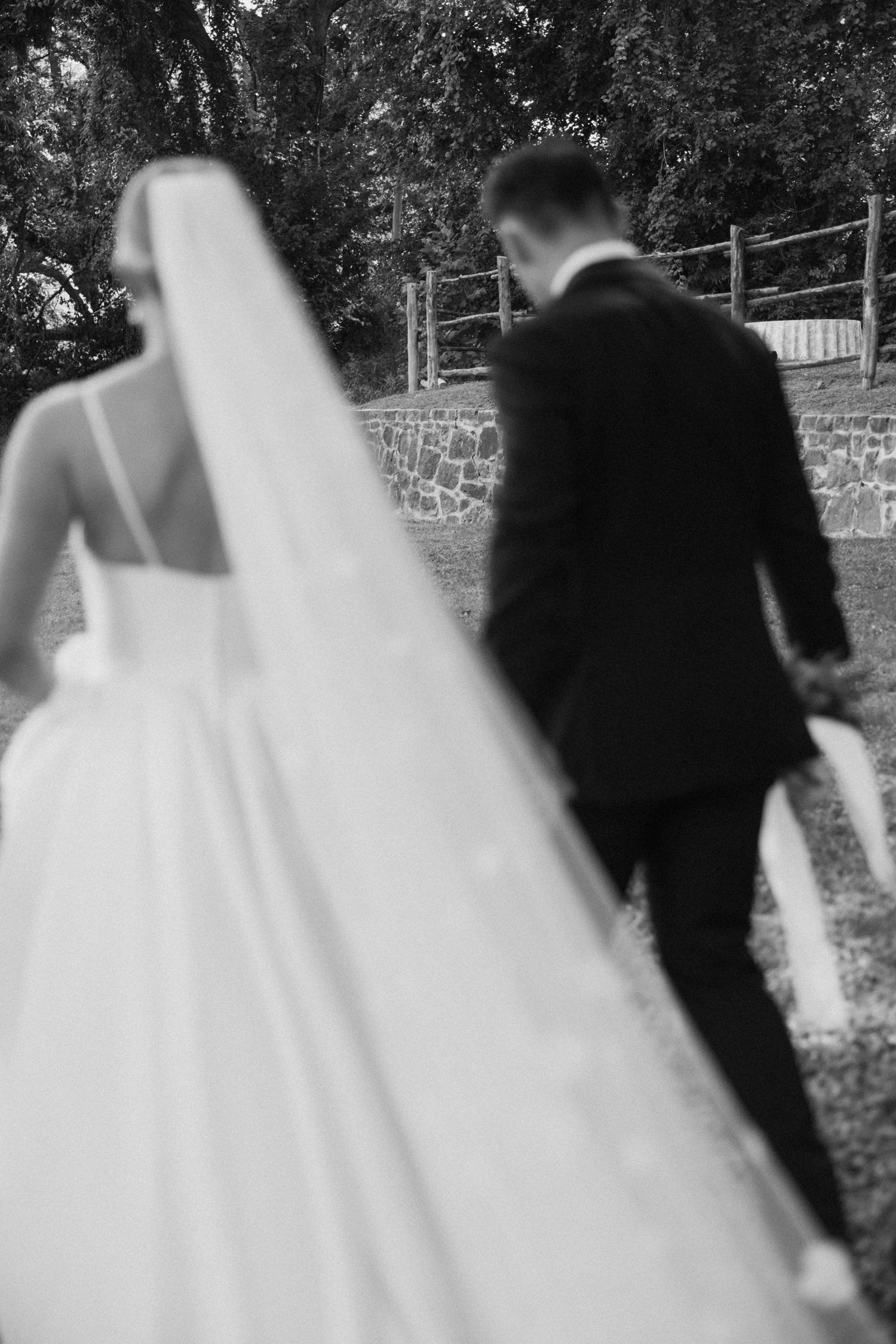 A black and white wedding photo showing a bride in a flowing white dress and groom in dark suit walking together outdoors.