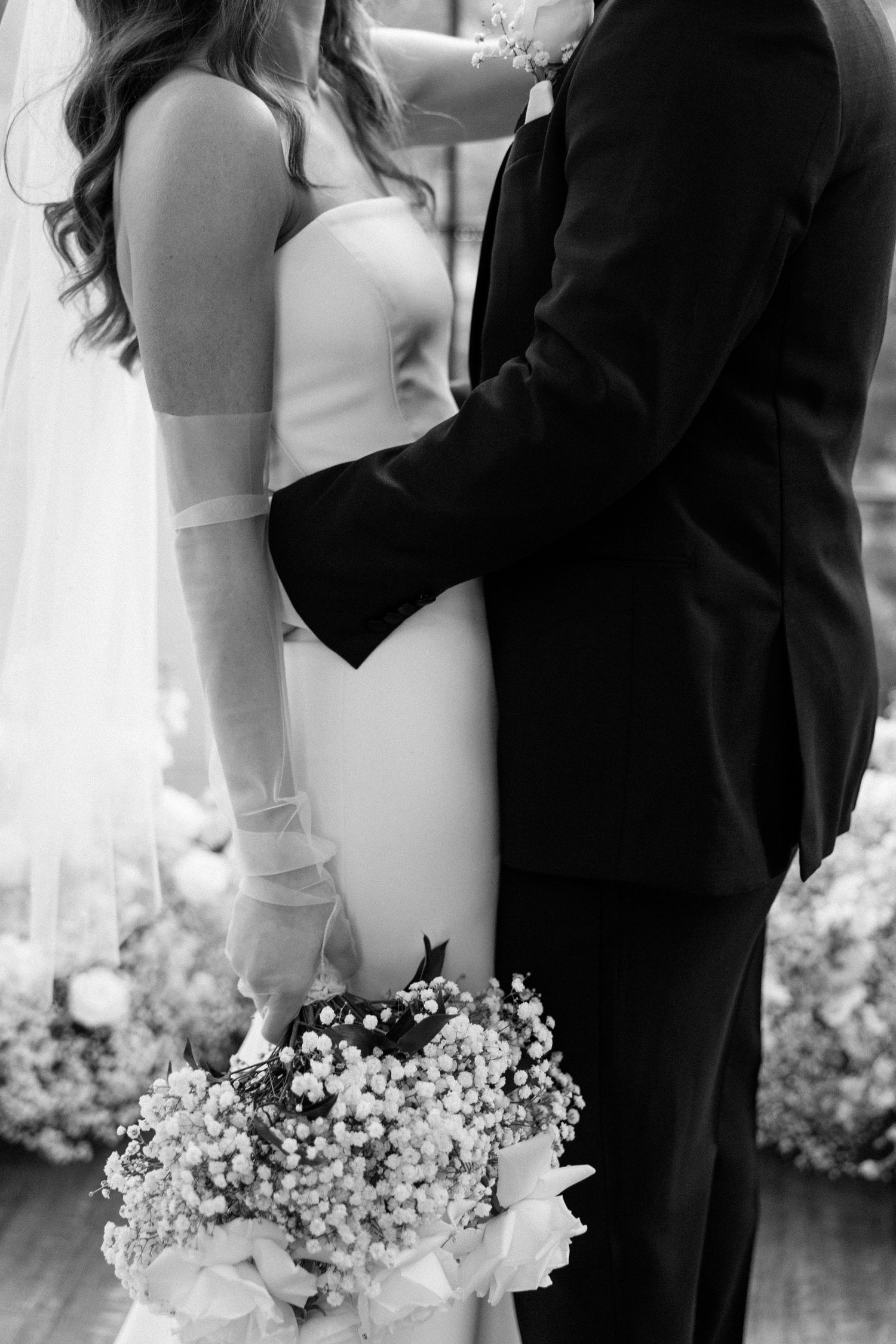 Black and white wedding photo showing close embrace of couple with bride holding delicate baby's breath bouquet.