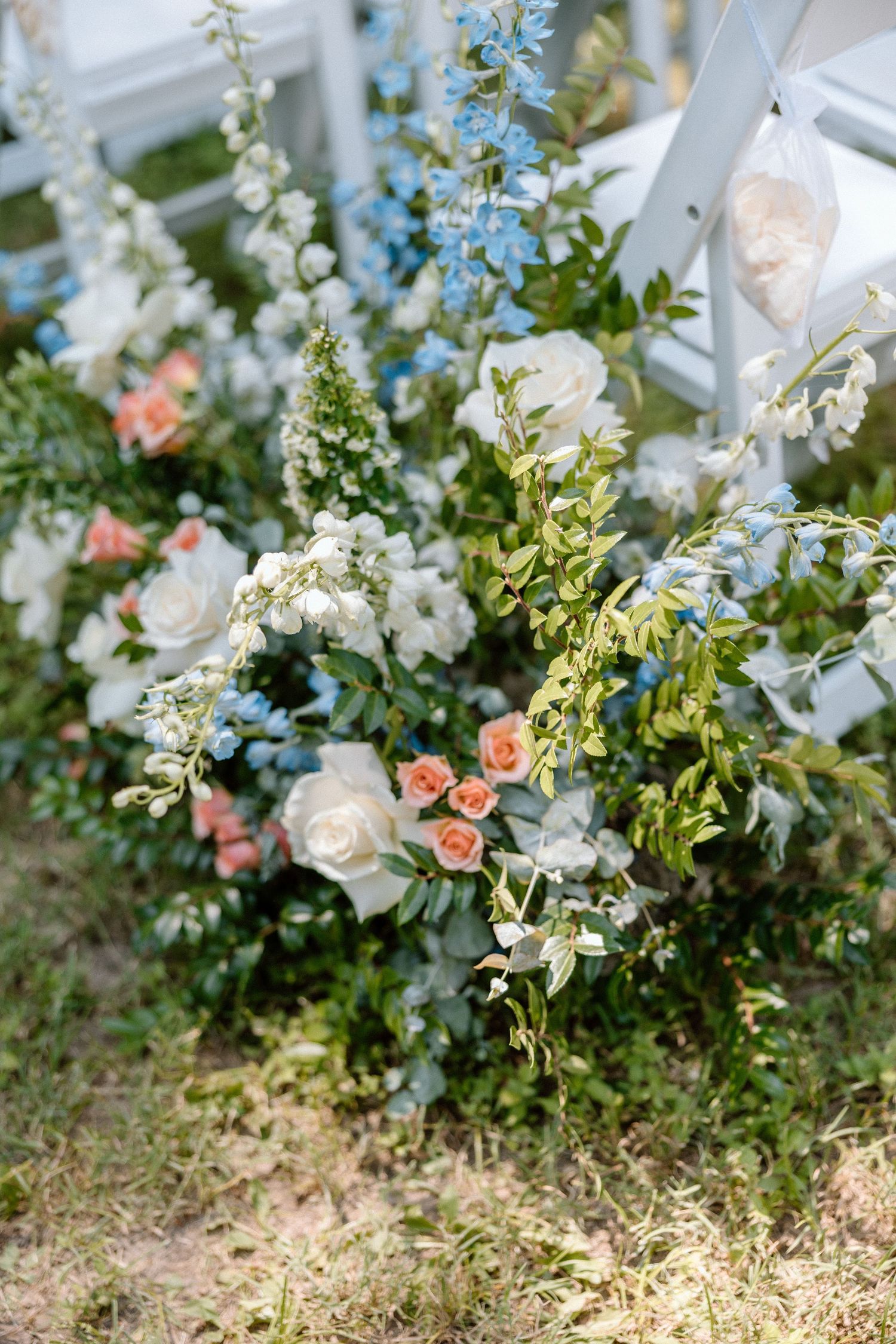 Delicate floral arrangement with white roses, peach spray roses, blue delphiniums and greenery decorating outdoor chairs.