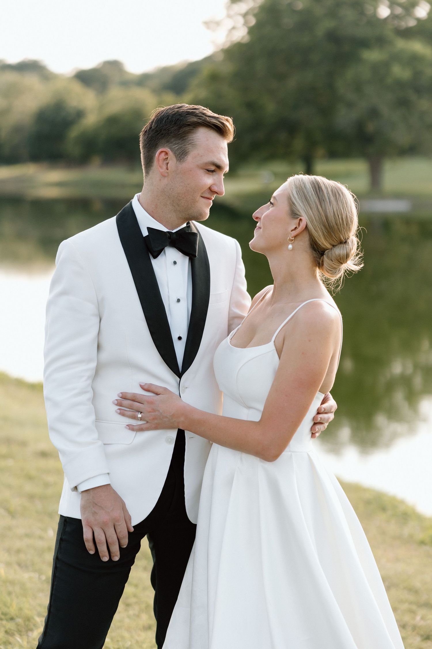 Newlywed couple shares intimate moments by a lake during sunset in formal wedding attire.