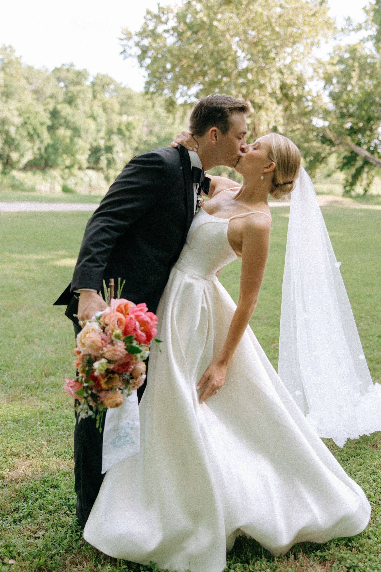 A romantic wedding couple in black and white attire share a kiss outdoors in a grassy field with flowing veil and flowers.