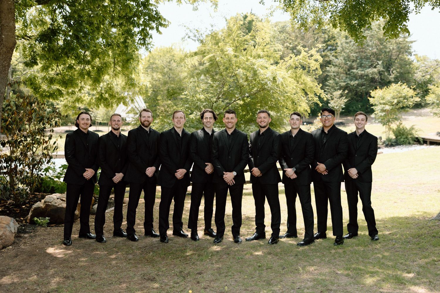 Wedding party in black formal attire stands in line outdoors against green foliage background.