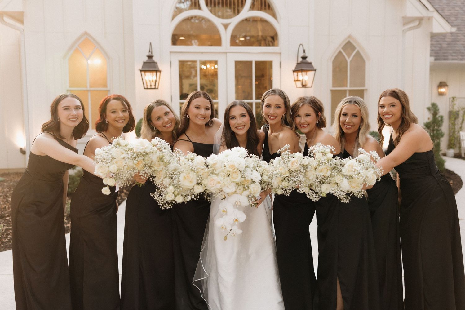 Wedding party in black dresses poses with white floral bouquets against a light colored wall.