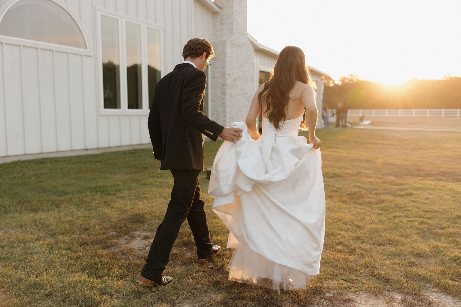 A couple in wedding attire holds hands and walks together at sunset outside a rustic white barn.